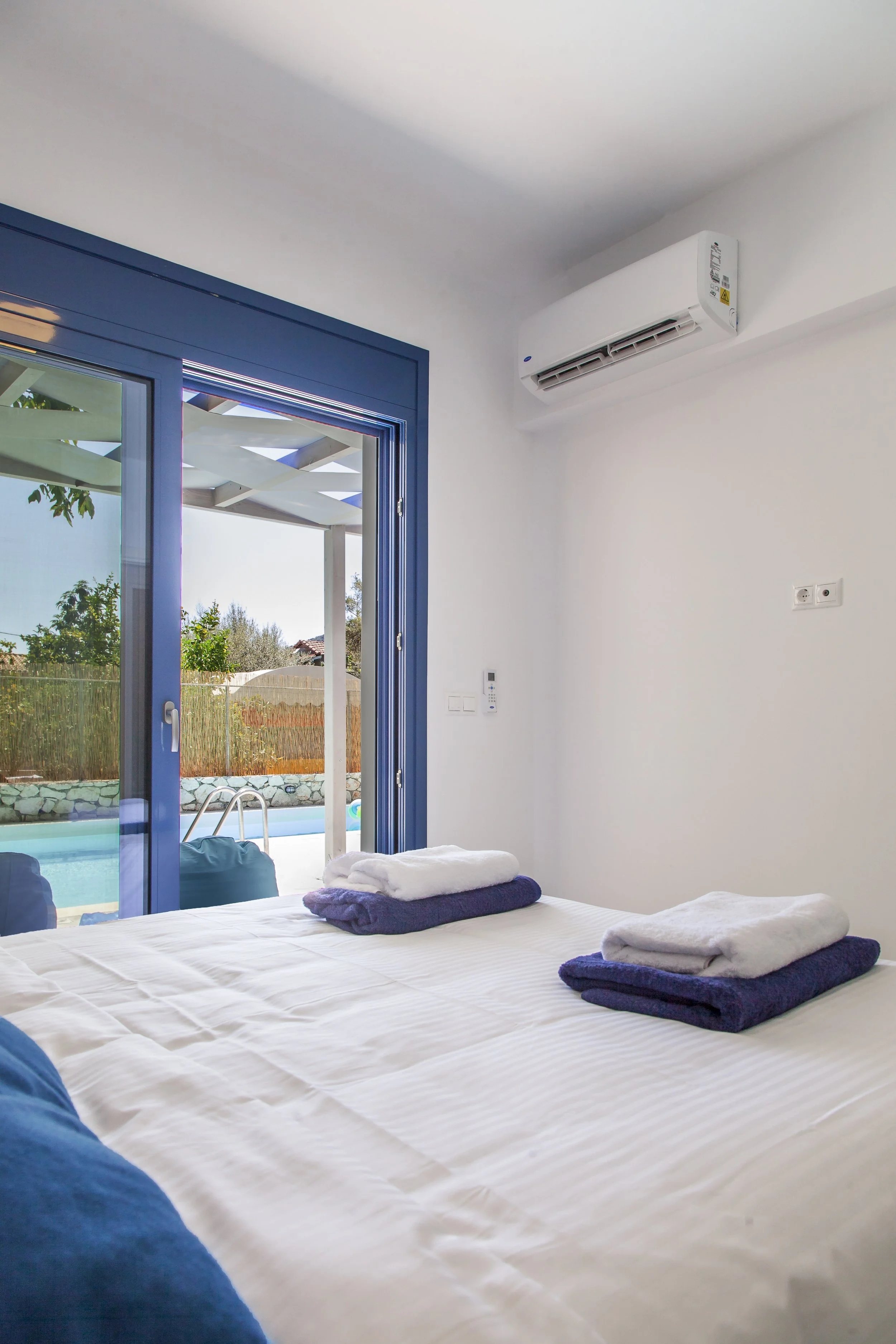 A bedroom with a large bed covered in white sheets and folded towels, large blue sliding glass doors leading to a backyard with a pool, trees, and a pergola, and white walls with a wall-mounted air conditioner.