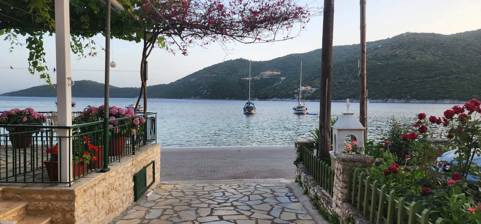 A waterfront scene with a paved stone path leading to a small dock. Potted flowers and bushes with pink and red blossoms are on both sides. Sailboats are anchored on the water, with green hills and mountains in the background. A tree with pink flower