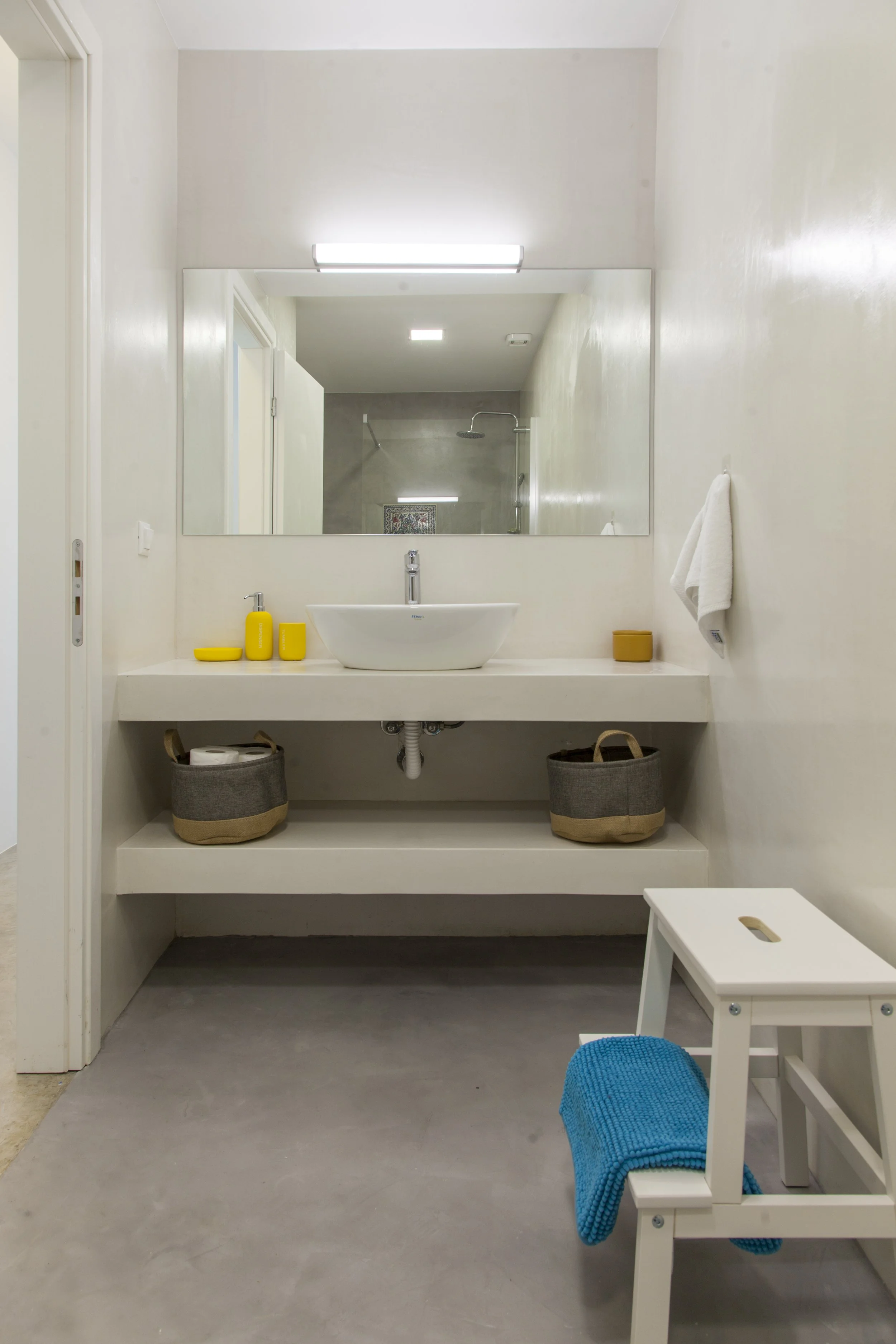 Minimalist bathroom with white walls, a large mirror, and a white vessel sink. Yellow toiletries on the counter, a white towel hanging on the wall, and baskets underneath. Small white stool with a blue blanket, in front of a white chair with a blue c