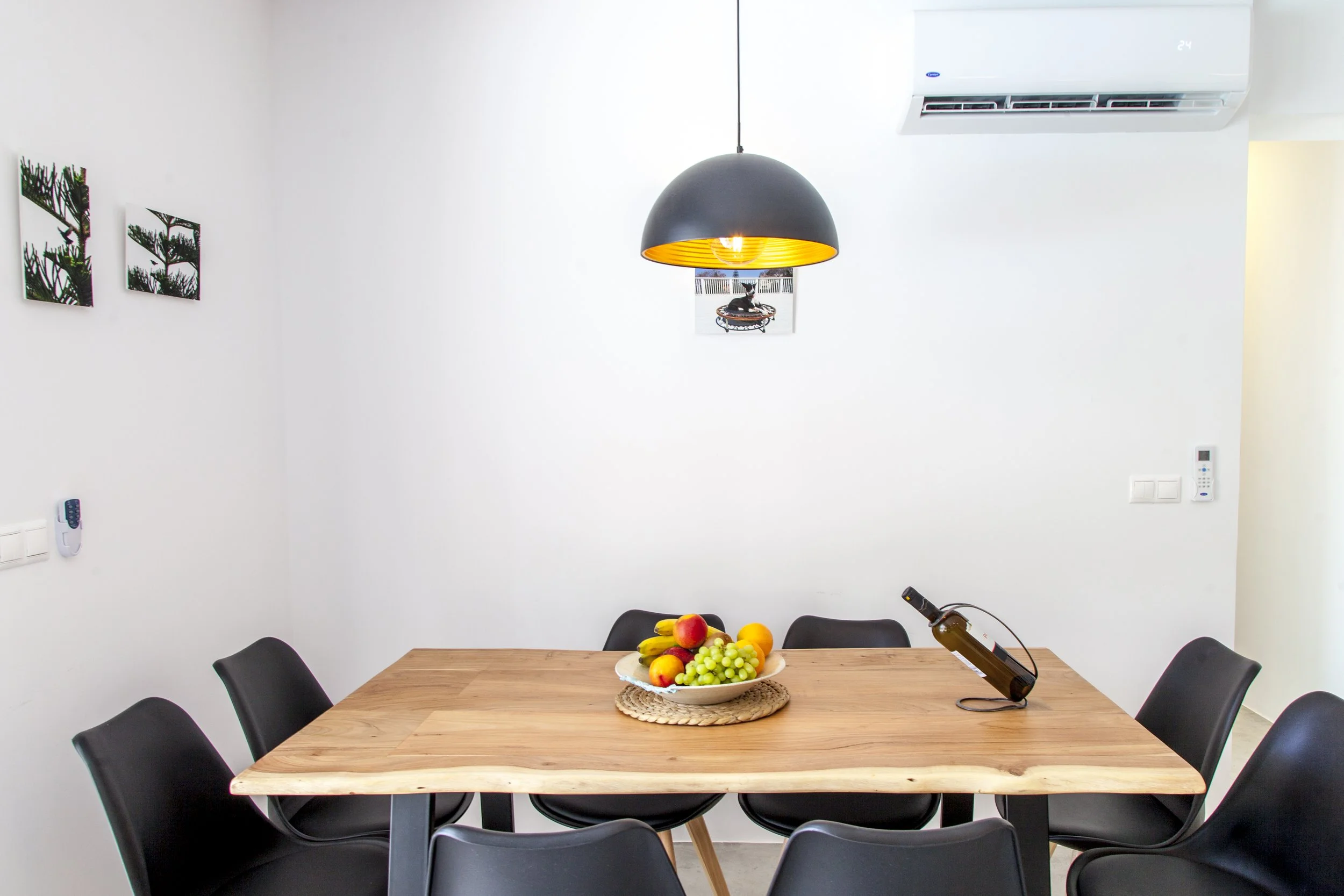 A dining room with a wooden table, six black chairs, a bowl of assorted fruits, and a wine bottle. The background features plain white walls, a hanging black pendant light with a yellow interior, a small photo of a dog on a trampoline, an air conditi