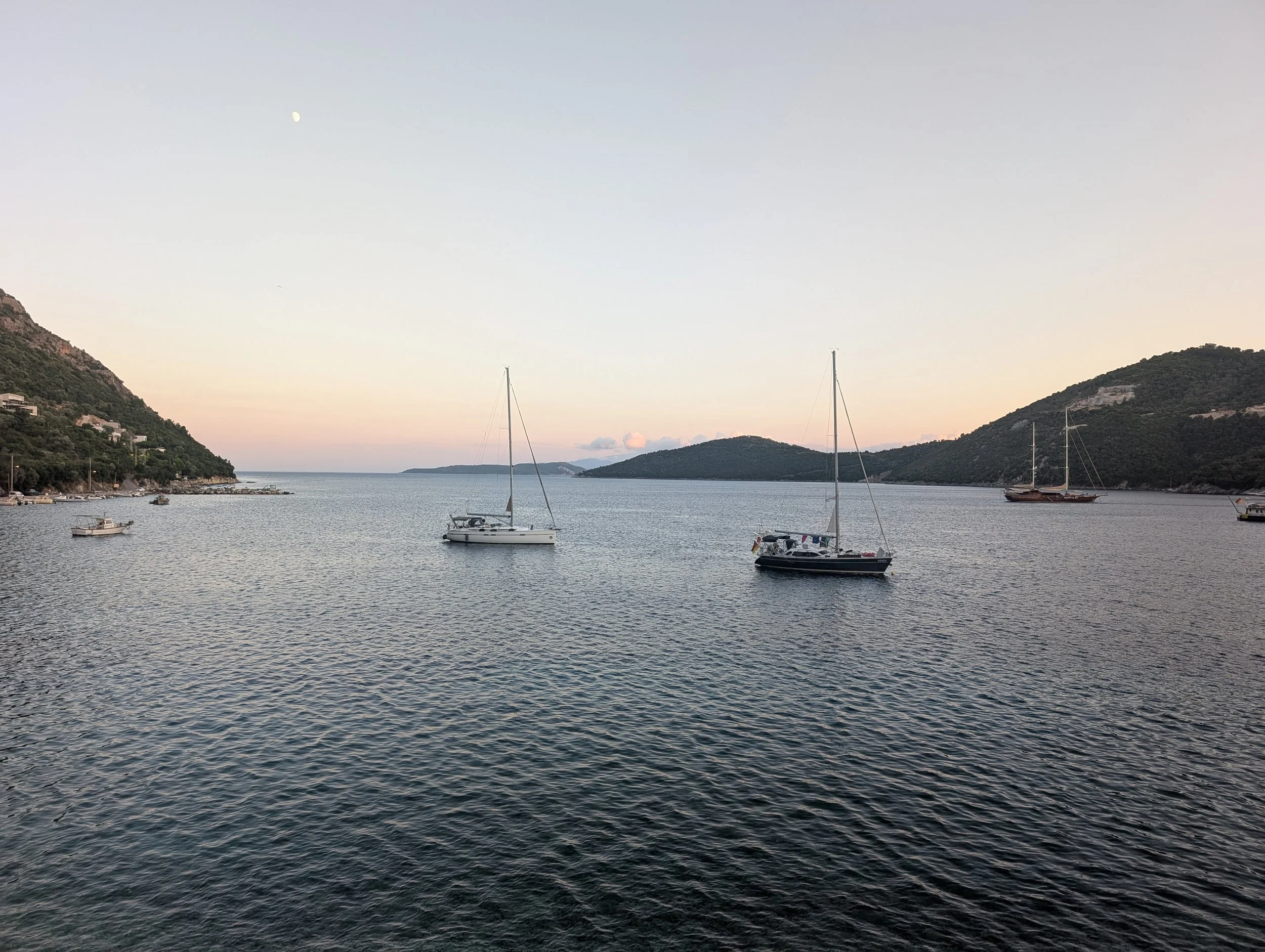 A calm body of water with sailboats and a small motorboat, surrounded by green hills, during the early evening with a partly visible moon in the sky.