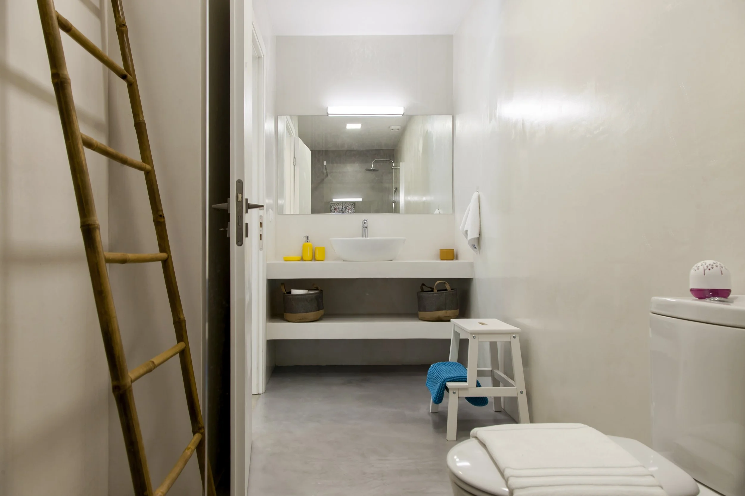 A minimalist bathroom with a white wall, a small wooden stool, and a toilet with a folded towel on top. There is a ladder on the left and a large mirror above a white sink with yellow toiletries. The shower area is visible in the reflection of the mi