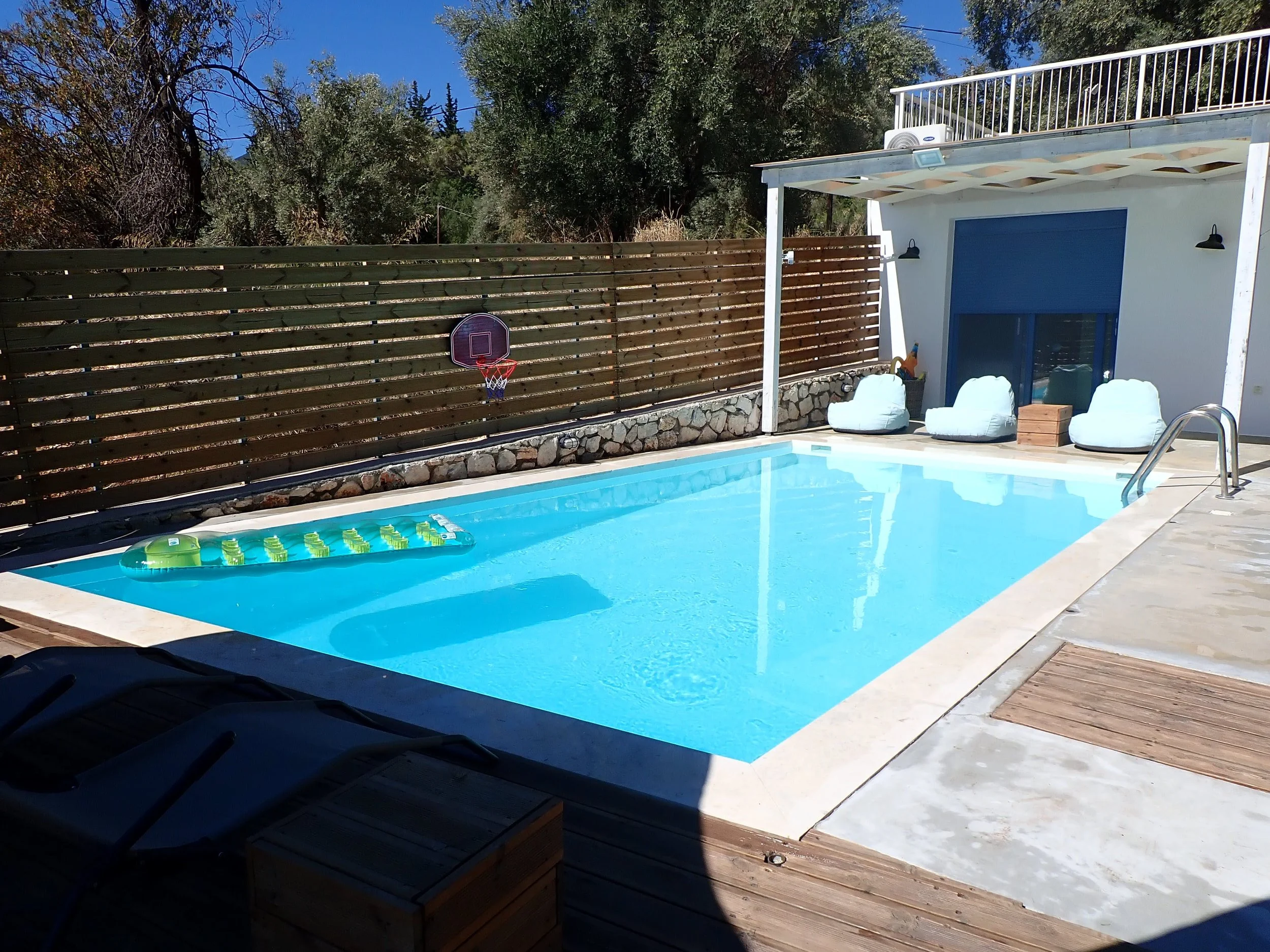 An outdoor backyard area with a swimming pool, a wooden fence with a small basketball hoop, and a patio with lounge chairs and bean bags. There are trees in the background and a two-story house structure with a balcony.