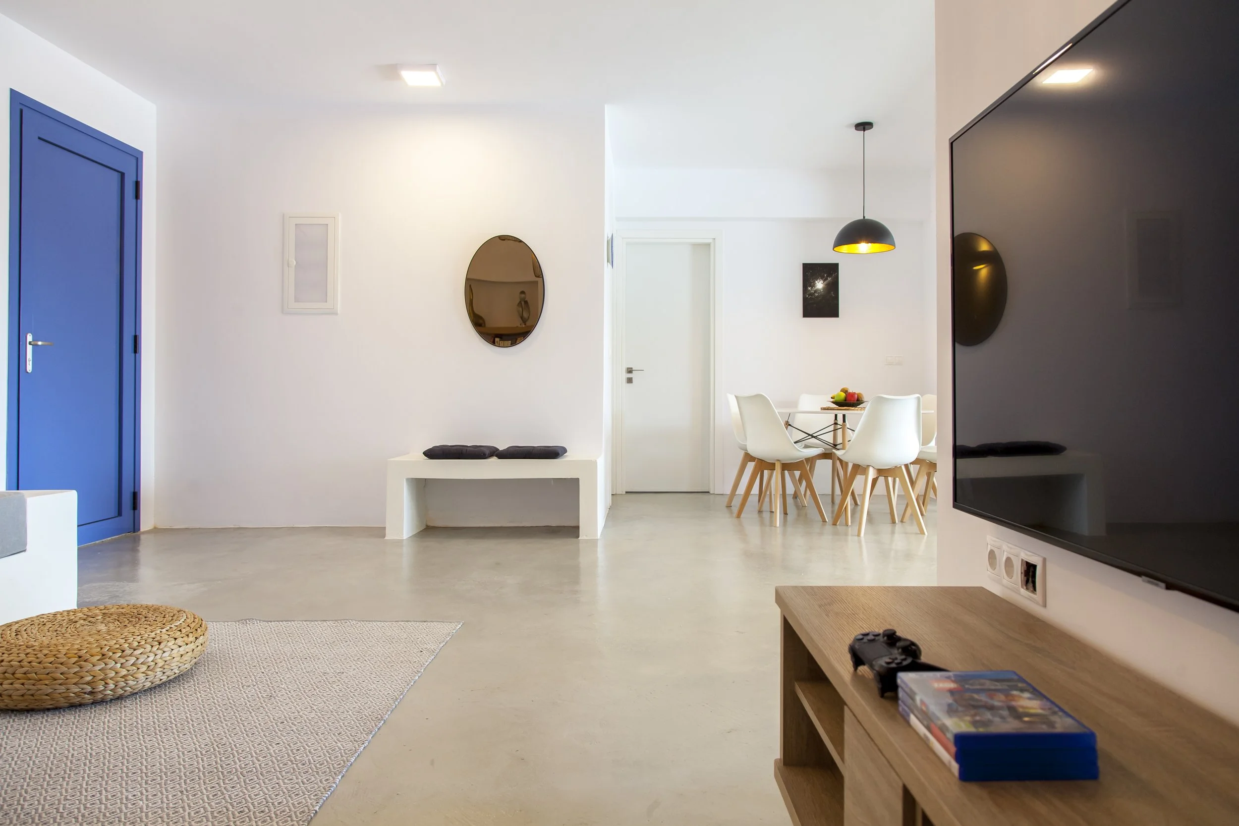 A modern, minimalist living space with a white wall, a blue door, a round mirror, a white bench with black cushions, a dining area with a black pendant lamp, white chairs, a wooden table with a fruit bowl, a black television, and a puzzle game on a w