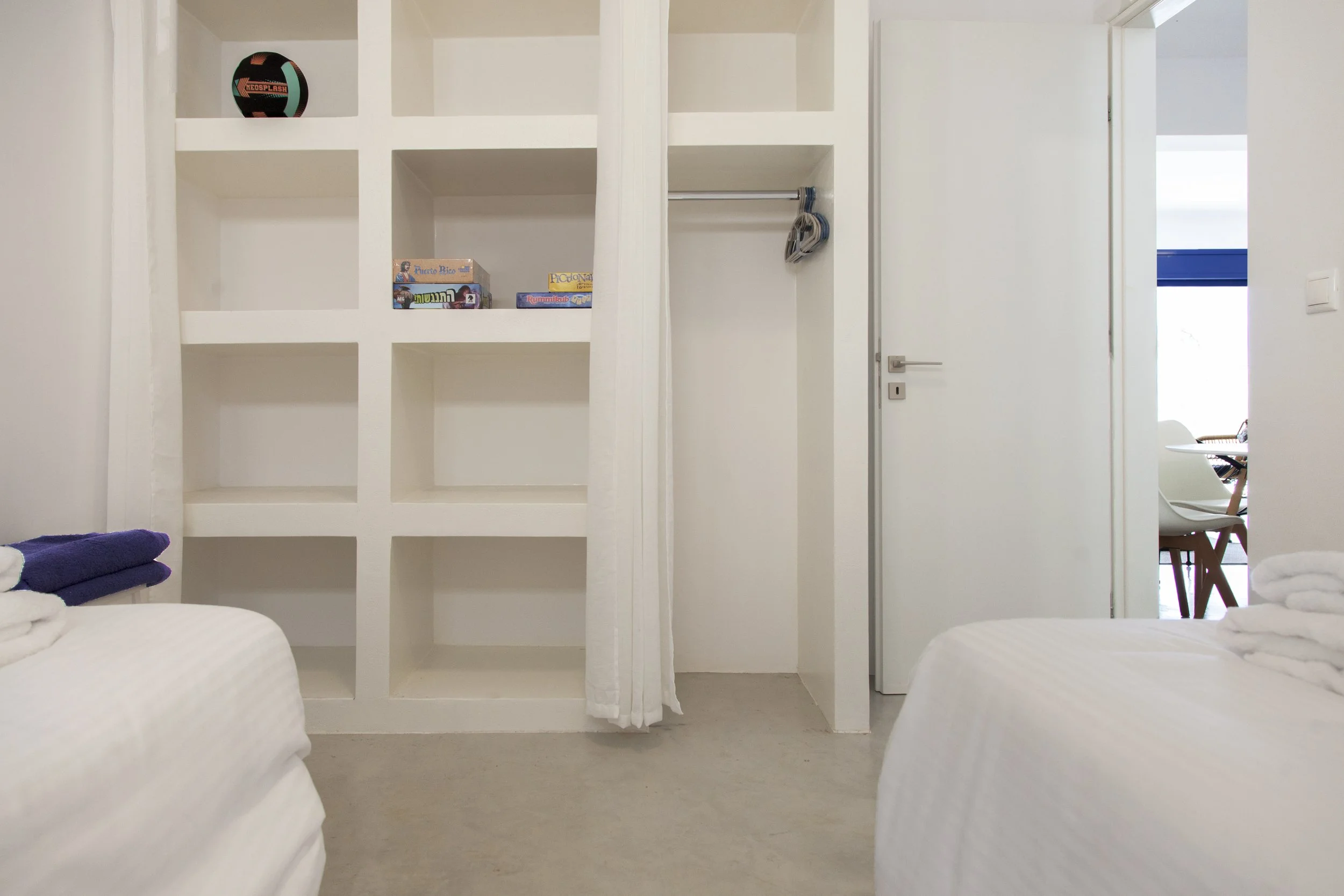View of a bright, minimalist room divider or wall with built-in shelves and a door, overlooking a living space with a blue wall and furniture in the background. Part of a bed with white linens and folded towels is visible in the foreground.