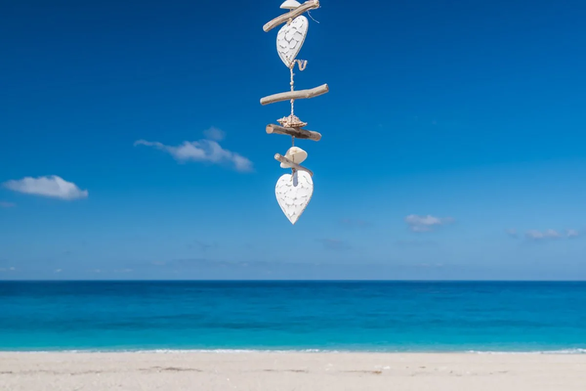 A hanging decoration with wooden hearts and fish shapes on a beach with the ocean and clear blue sky in the background.