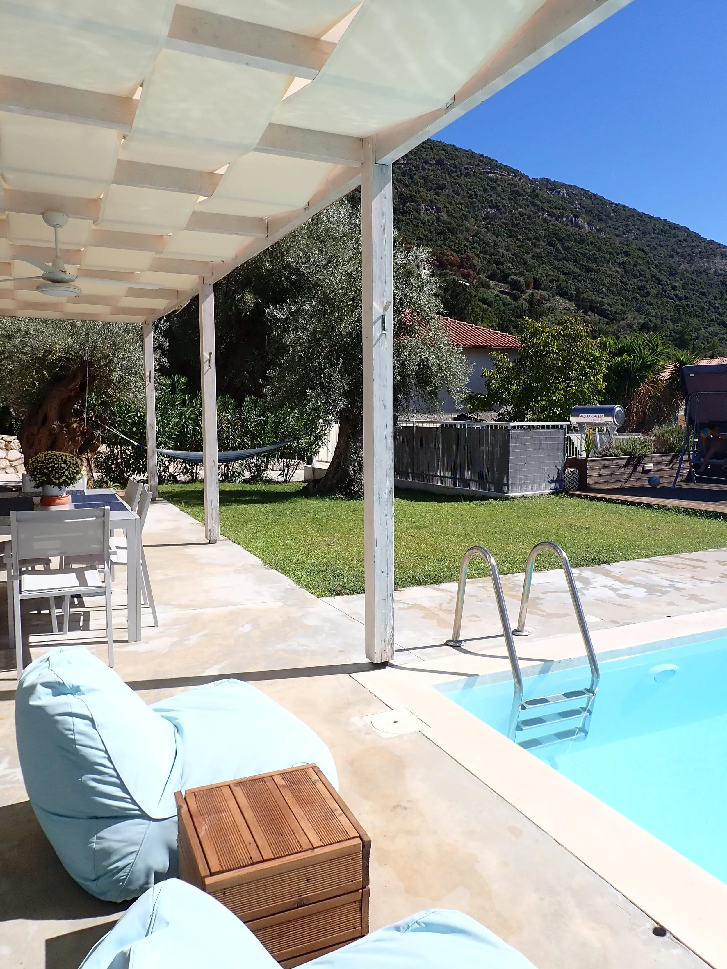 View of a backyard with a swimming pool, lounge chairs, a white pergola with a ceiling fan, surrounded by green trees and hills in the background, under a clear blue sky.
