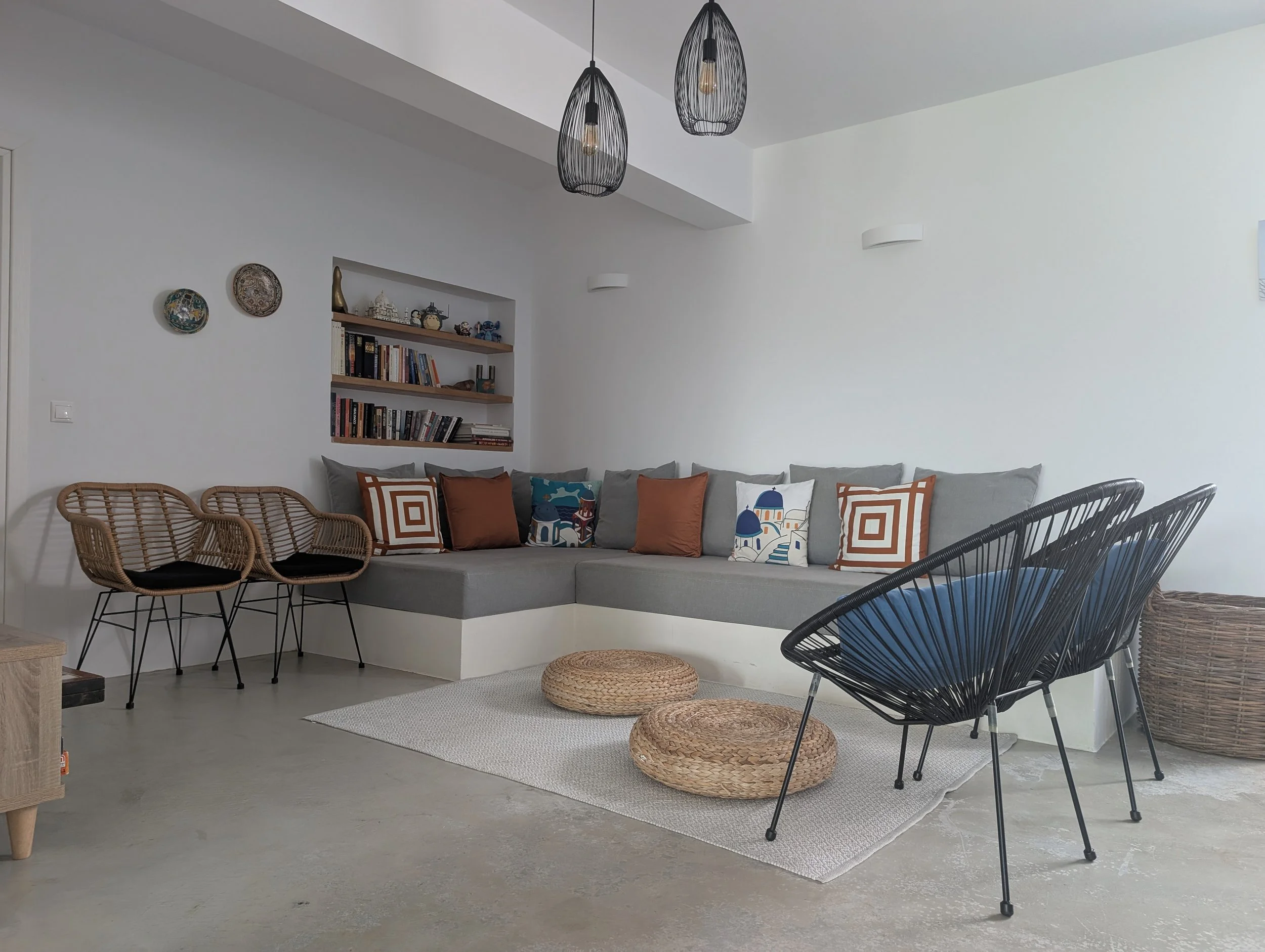 Living room with a gray sectional sofa decorated with colorful throw pillows, a white textured area rug, wicker and black wire chairs, woven ottomans, a white wall with Asian decorative plates, and a wooden bookshelf filled with books and ornaments. 