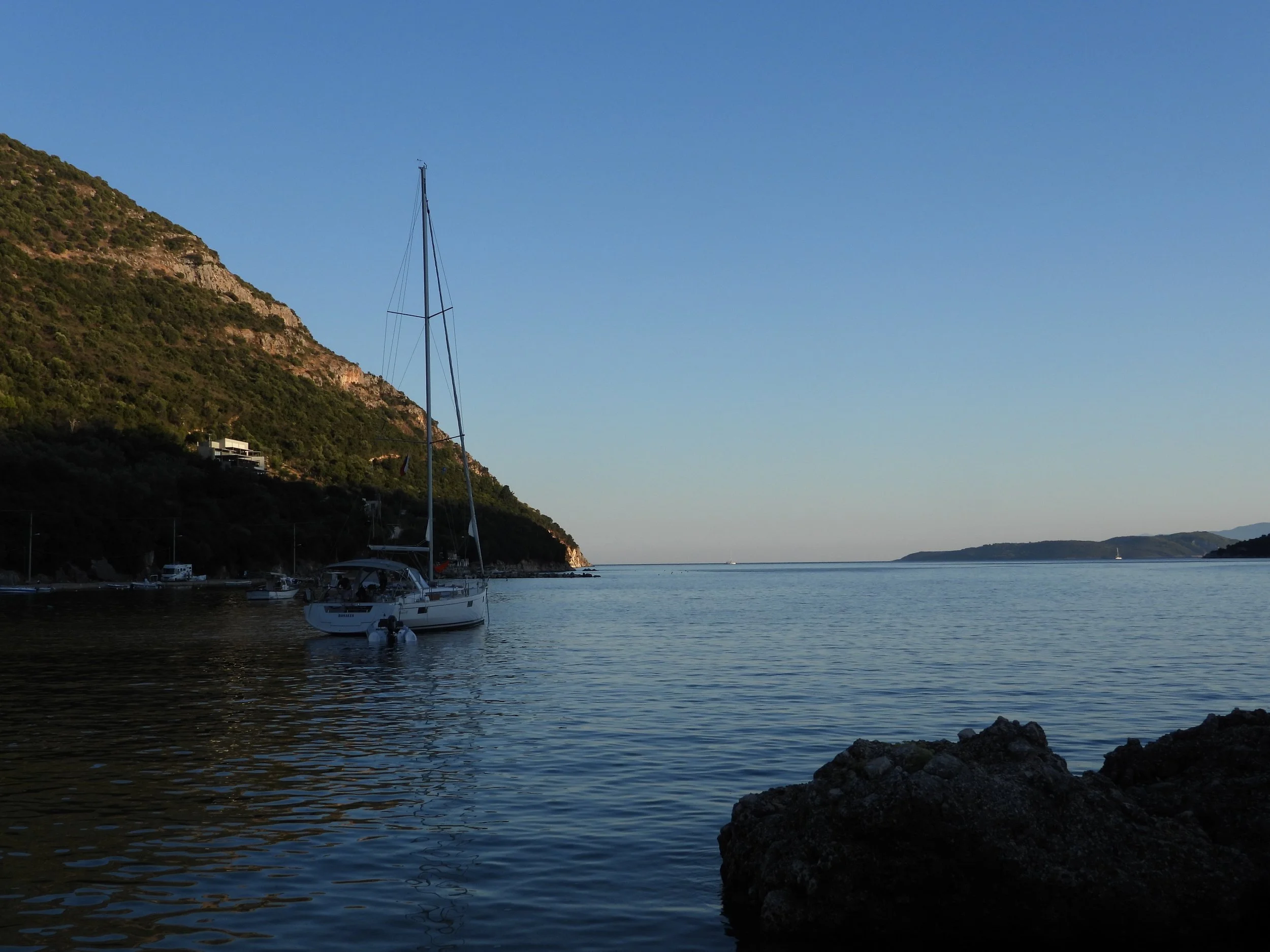 A peaceful harbor scene with boats docked along the shoreline, a hill covered with green vegetation on the left, and a calm body of water extending to distant hills under a clear blue sky.