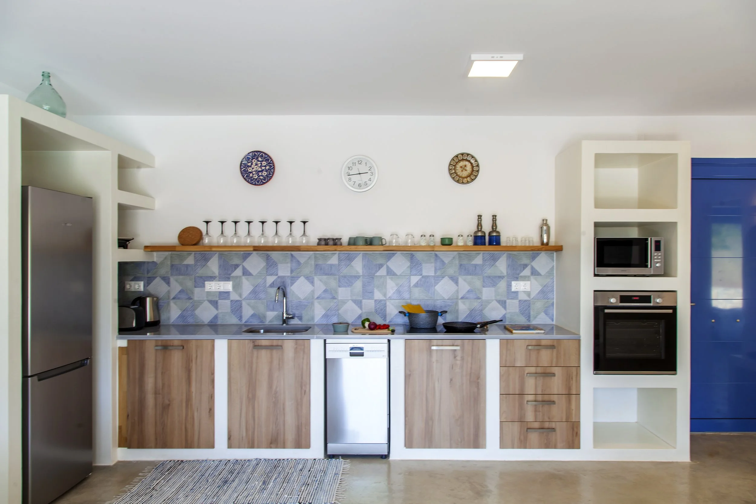 Modern kitchen with white walls, wooden lower cabinets, a gray and white geometric tile backsplash, stainless steel appliances including a refrigerator, microwave, and oven, a small kitchen island, and wall clocks. Decorative items and kitchenware ar