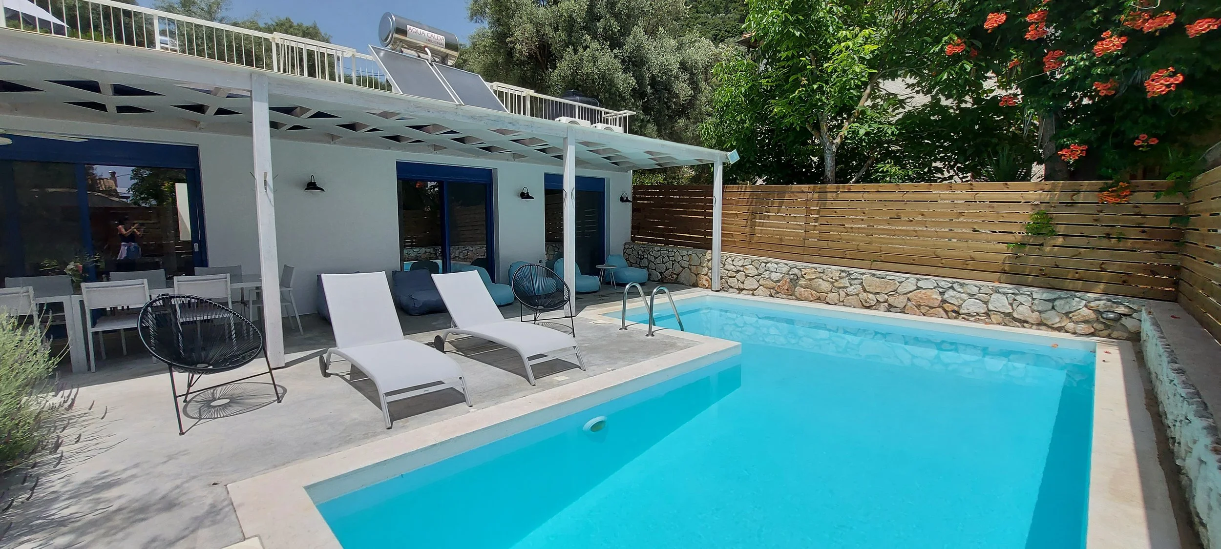 A backyard pool area with two white lounge chairs, a black circular chair, and cushions on the patio. There is a dining table with white chairs, a white wall with three glass doors, a wooden fence with a stone base, and lush trees and bushes in the background.