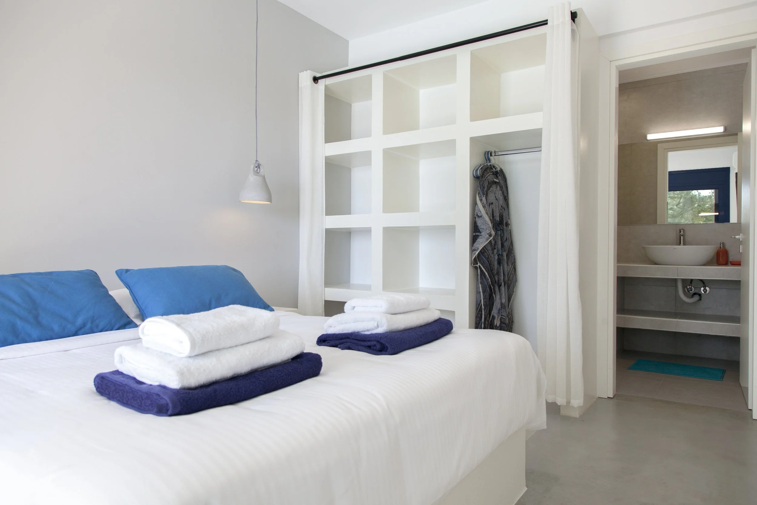 A bedroom with a large bed featuring blue pillows and folded towels, an open wardrobe with hangers and a curtain, and a bathroom with a sink and window in the background.