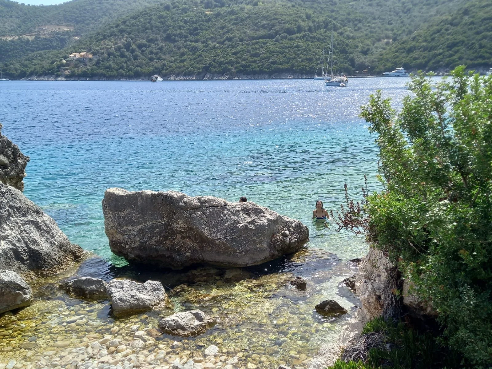 People enjoying a swim in a rocky, clear blue seaside cove with boats anchored across the water and lush green hills in the background.
