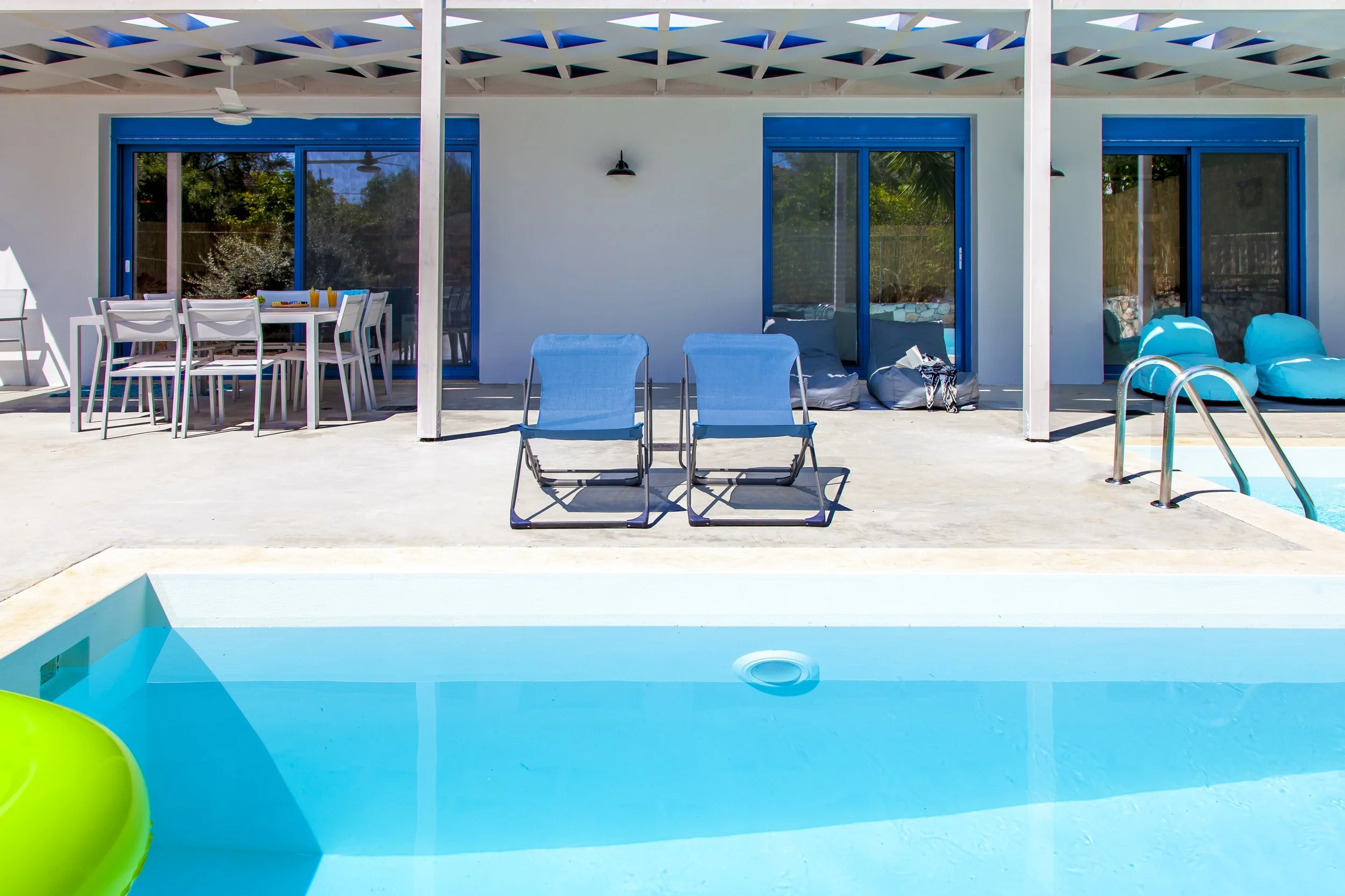 View of a swimming pool patio with blue lounge chairs, an outdoor dining table with white chairs, and sliding glass doors leading into a house, with a white pergola overhead.