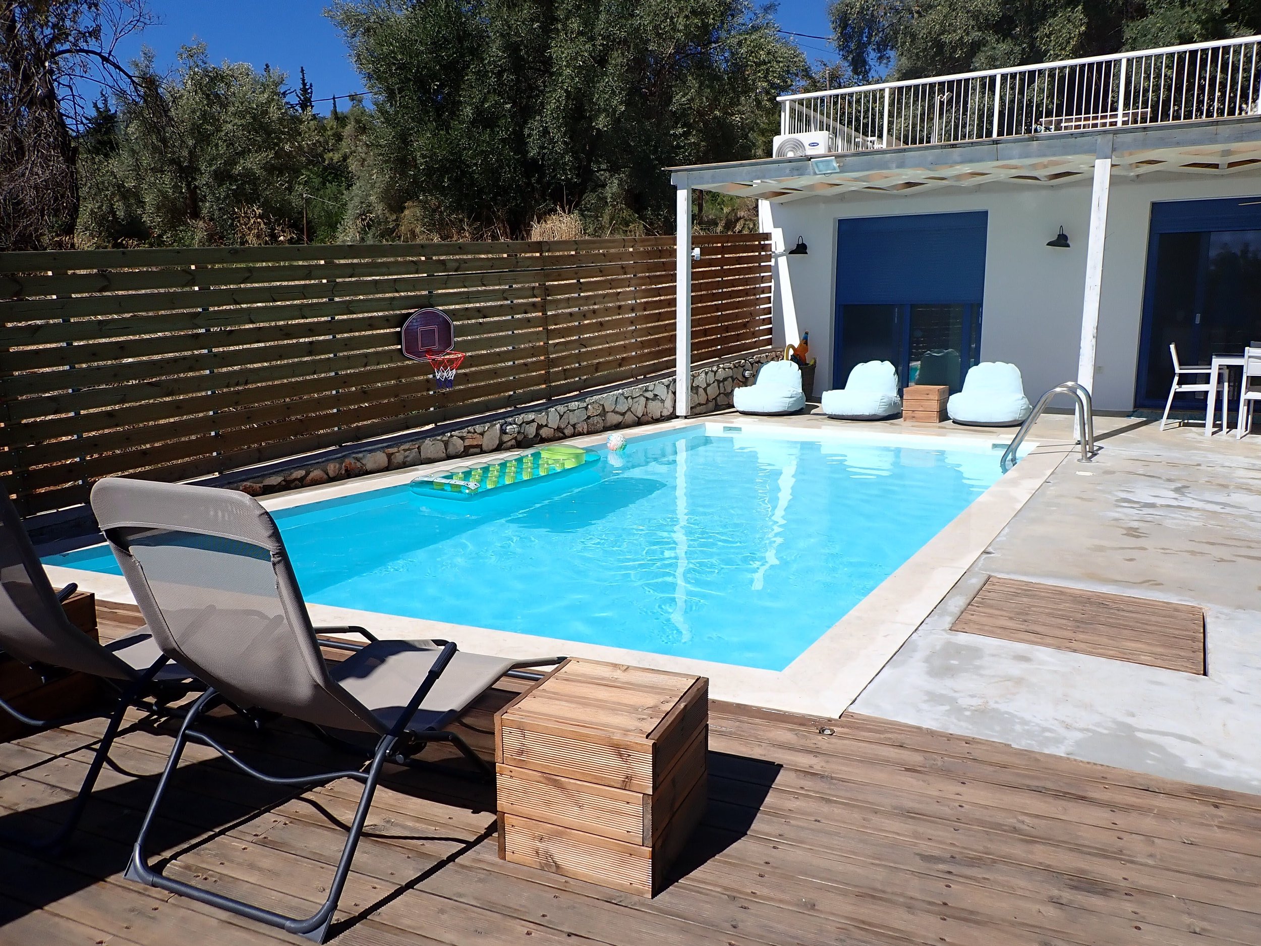 A backyard with a swimming pool, poolside chairs, a wooden deck, a privacy fence, and a house with a balcony. There is a basketball hoop attached to the fence, and trees and clear blue sky in the background.