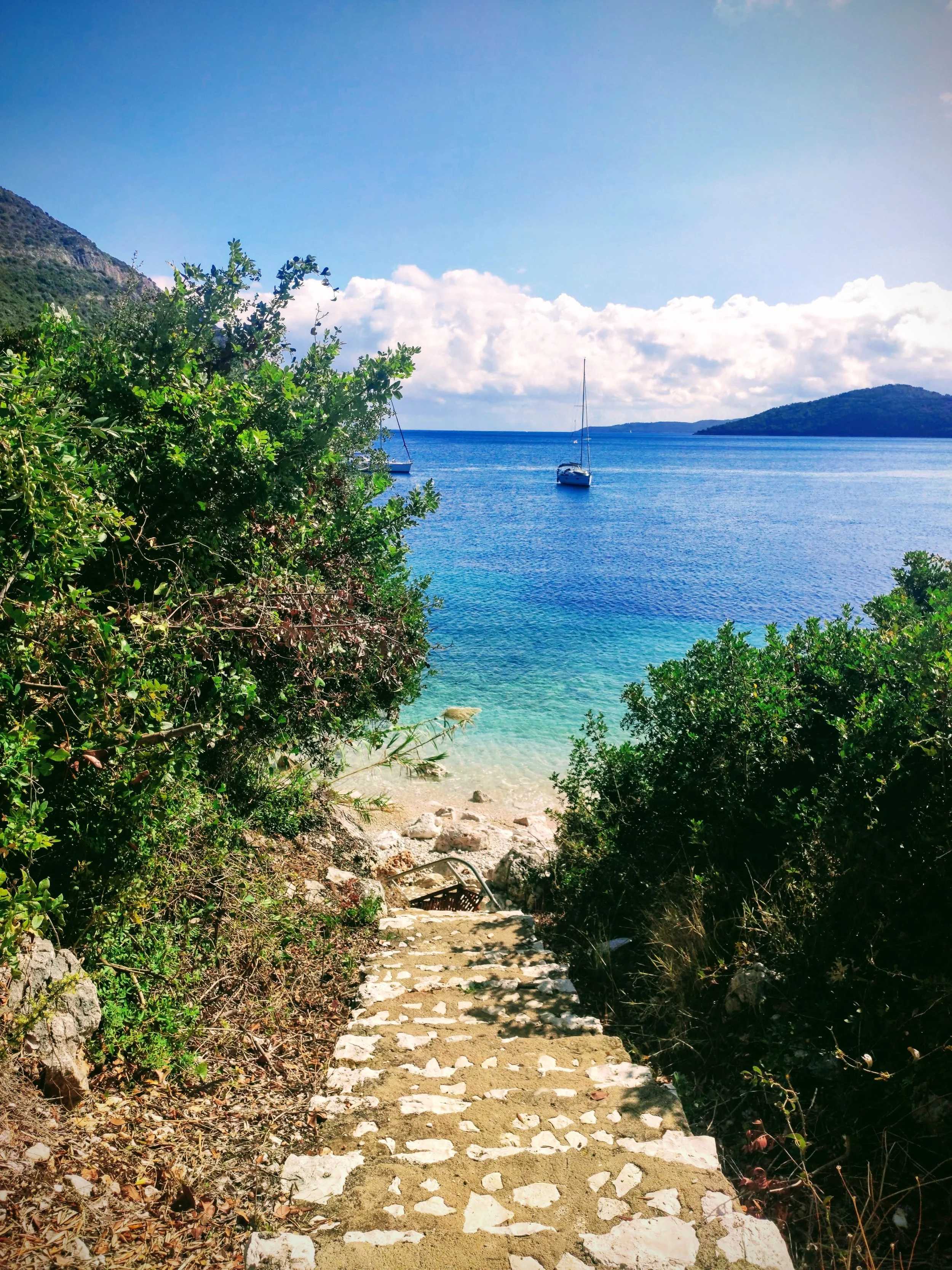 Photograph of a stone stairway leading down to a sandy beach with a turquoise ocean, two sailboats floating on the water, green bushes on either side of the stairs, a mountain slope on the left, and a distant island under a partly cloudy sky.
