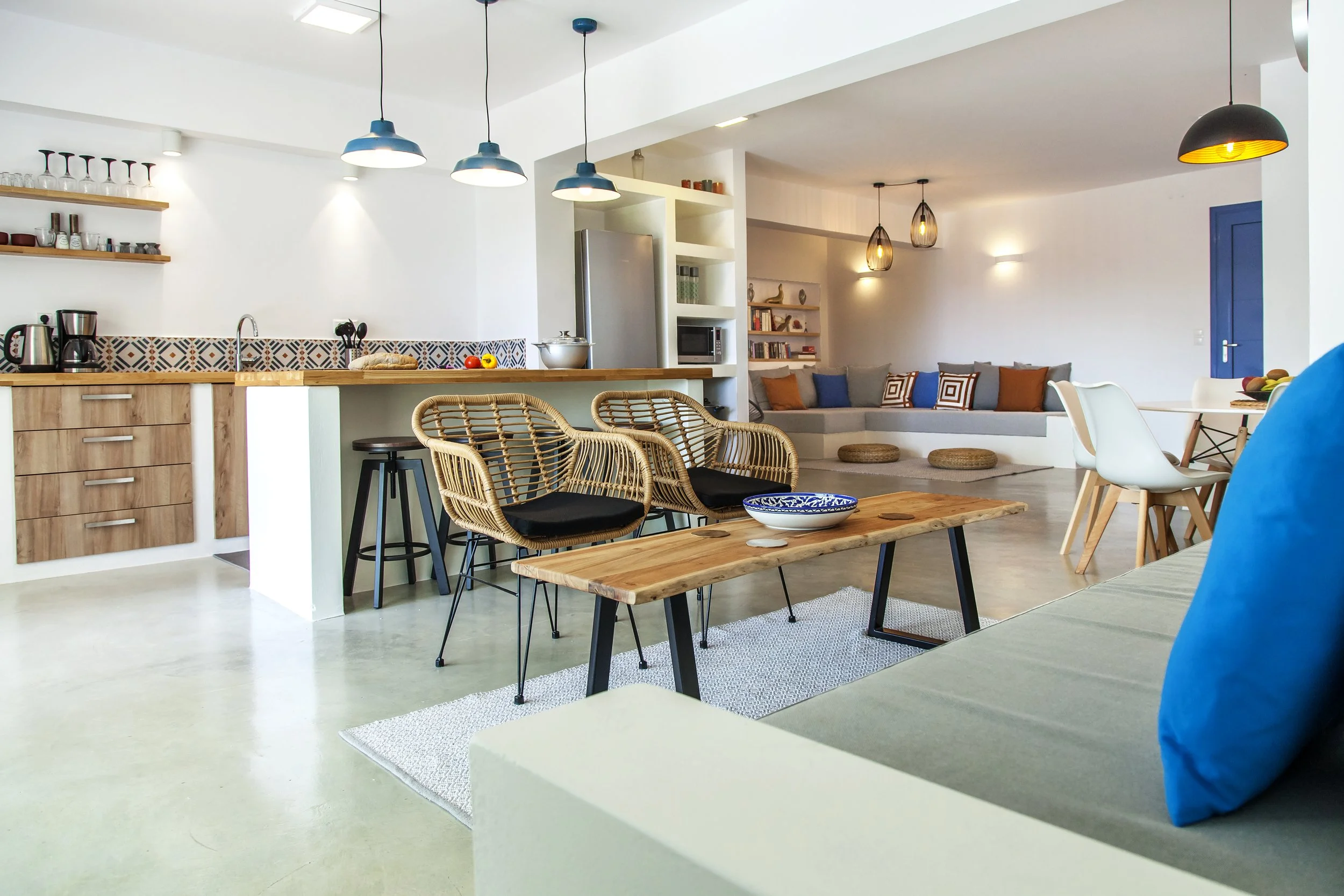 Open-concept living space with a kitchen, dining area, and lounge. White walls and ceiling, pendant lighting, wooden accents, and modern furniture with colorful cushions.