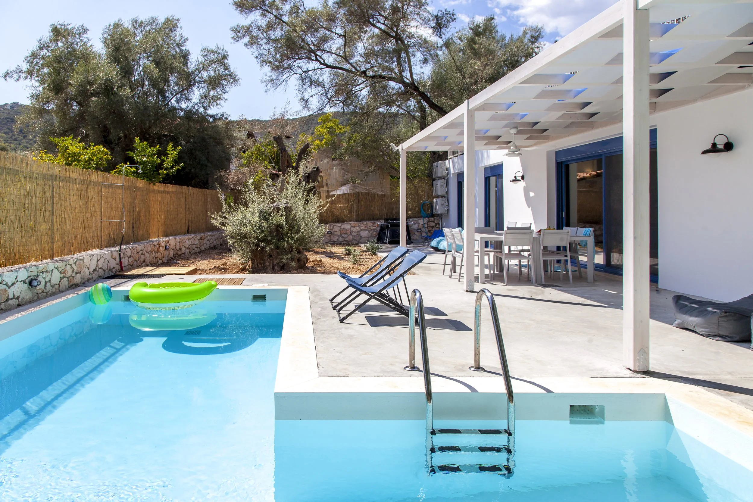 A backyard with a swimming pool, lounge chairs, a patio table with chairs, a fence along the yard, trees in the background, and a covered patio area with ceiling fans and outdoor lighting.