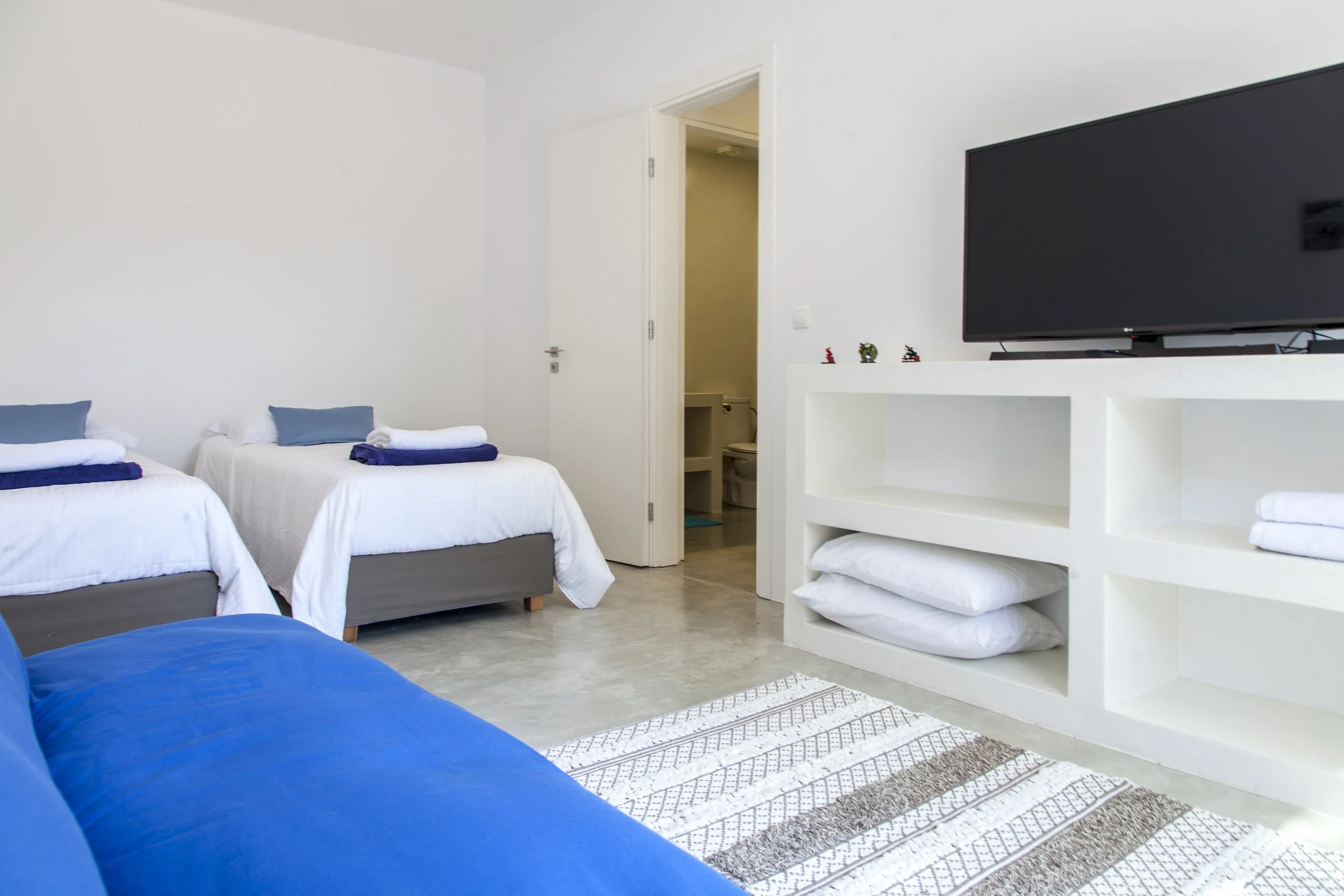 A brightly lit room with three beds, pillows, and towels, a flat-screen TV on a white shelf, and an open door leading to a bathroom.