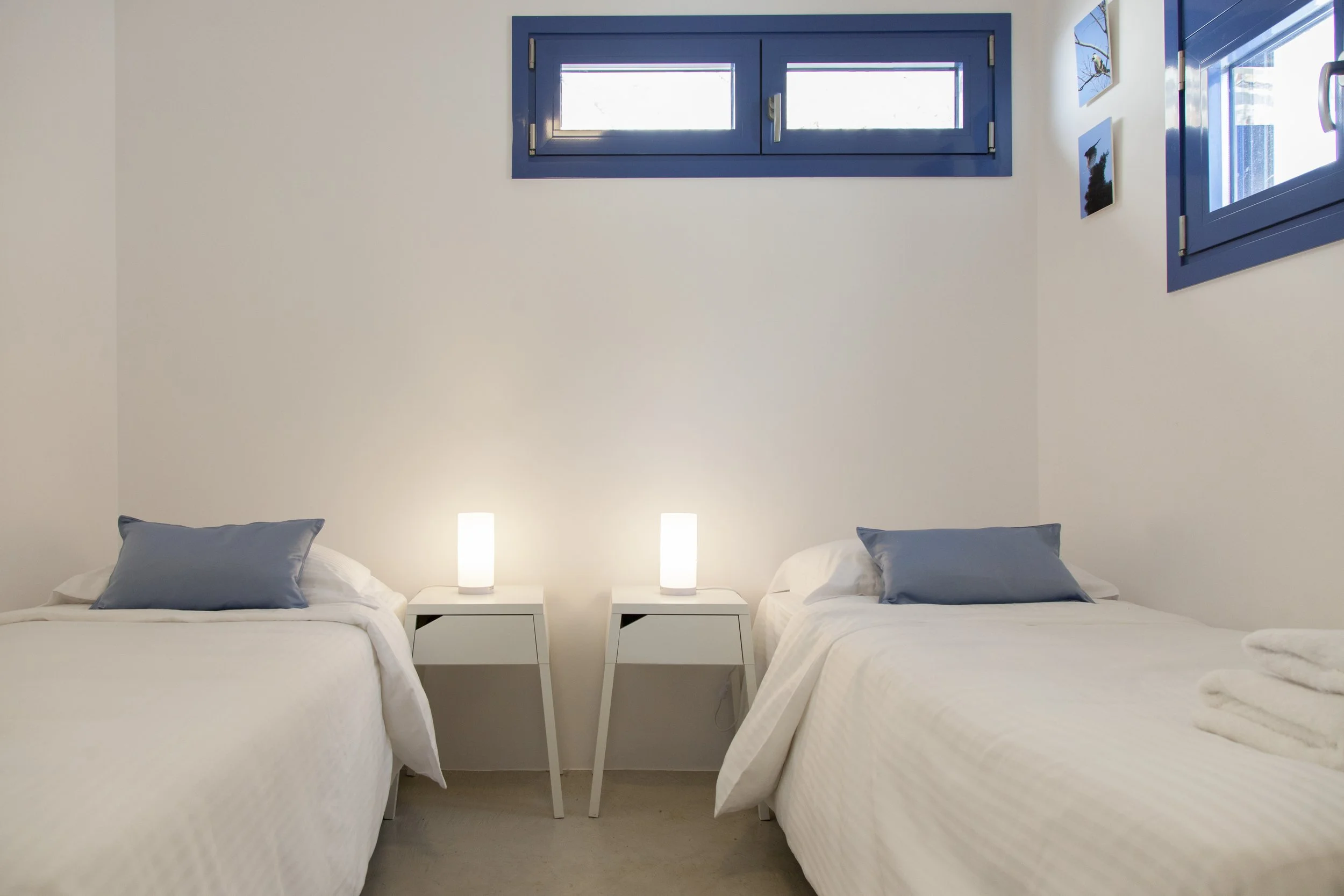 Two white beds with blue pillows, decorated with white bedspreads, separated by a small white nightstand with two modern white lamps, against a white wall with blue-framed windows and three framed pictures of birds.