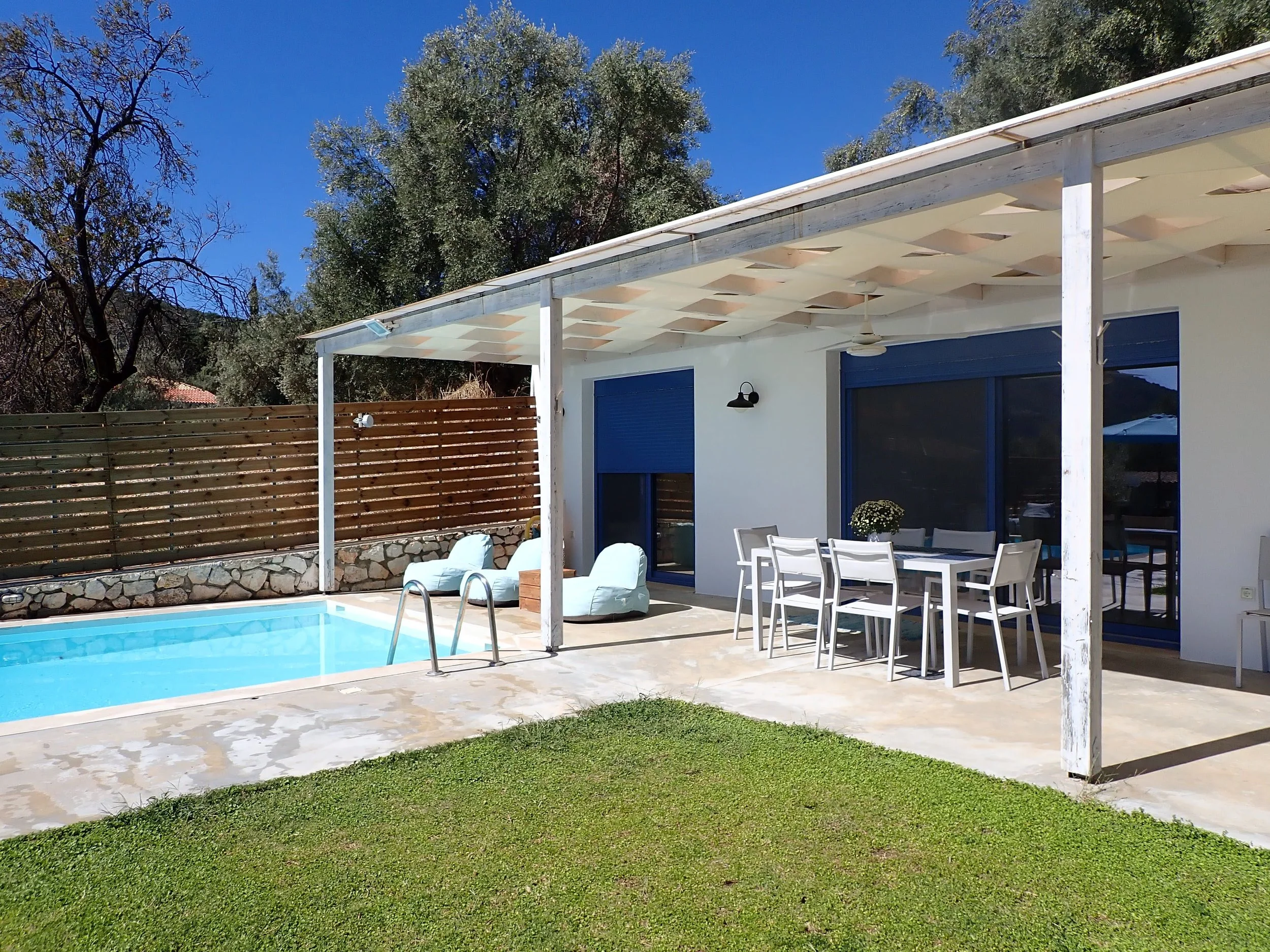 House with outdoor patio, pool, white outdoor furniture, and patio dining set, surrounded by green grass and trees under a clear blue sky.