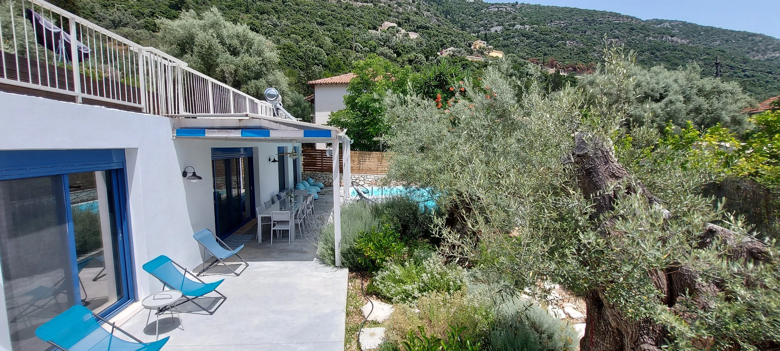 A house with a white exterior and blue window frames, located in a lush green hilly area. The house has a patio with lounge chairs, a dining table, and a yard with a swimming pool surrounded by trees and shrubs.