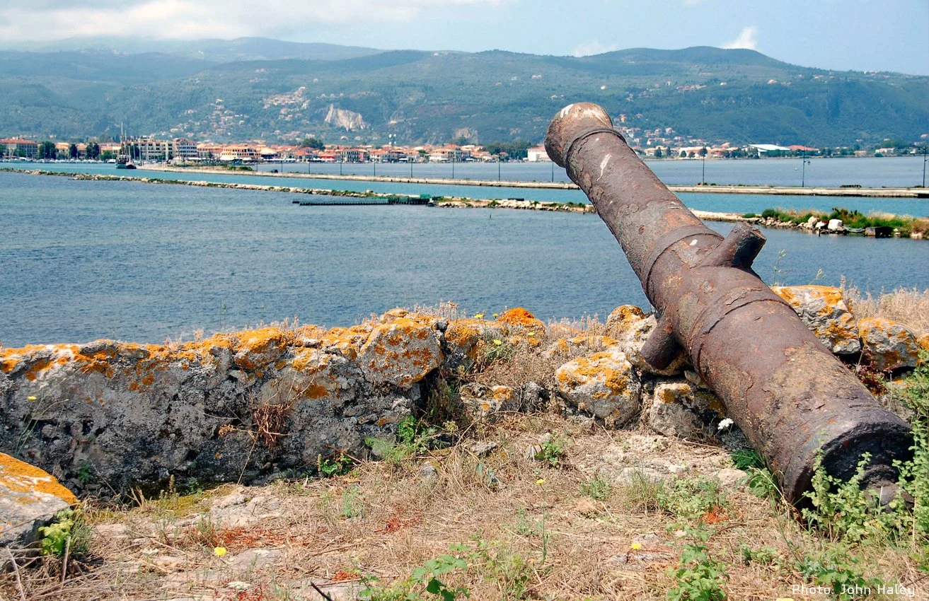 An old rusty cannon leaning on a stone wall overlooking a body of water with a town and mountains in the background.