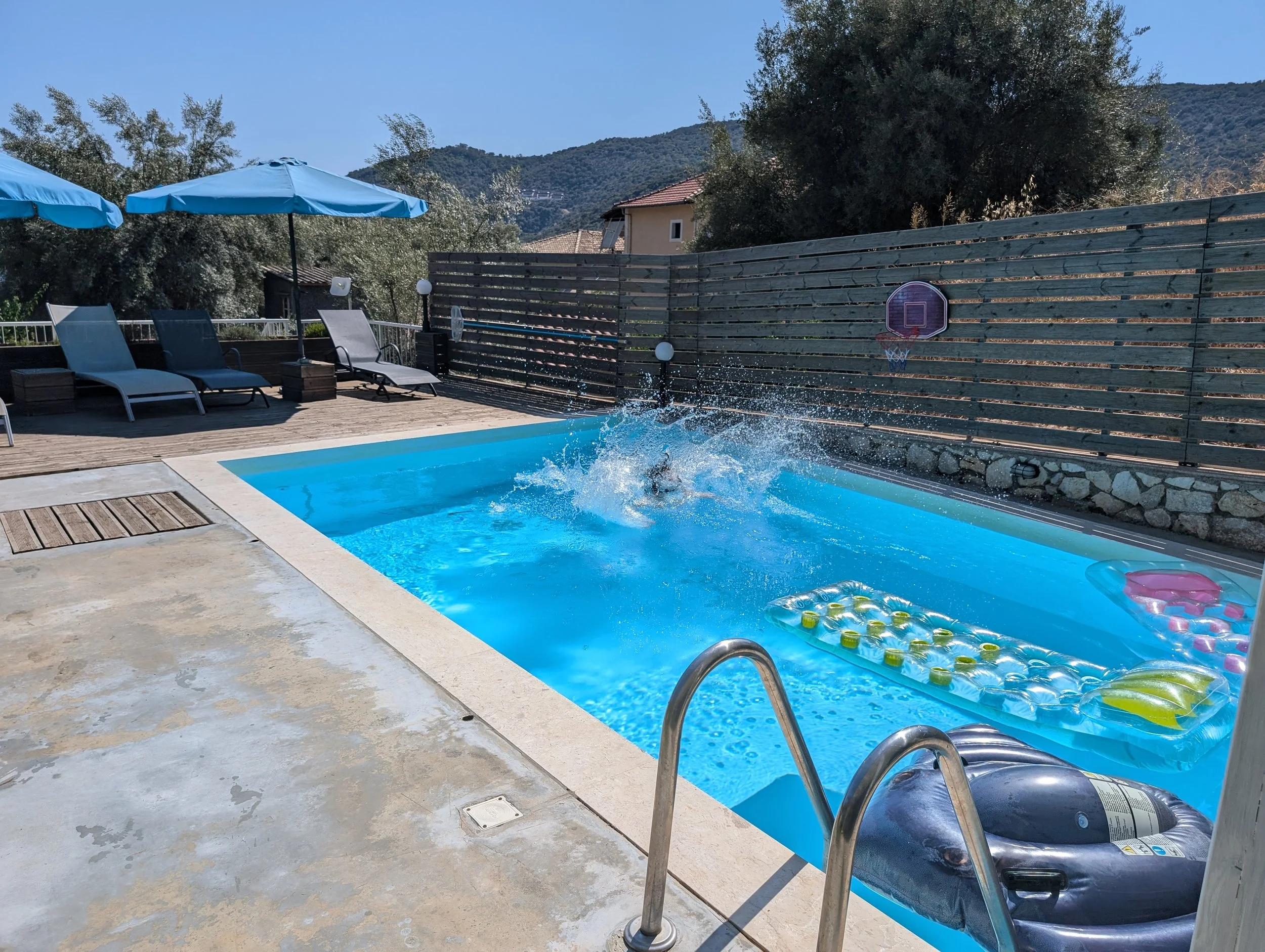 An outdoor swimming pool area with lounge chairs, blue umbrellas, a wooden privacy fence, a small basketball hoop, and a backdrop of trees and mountains.