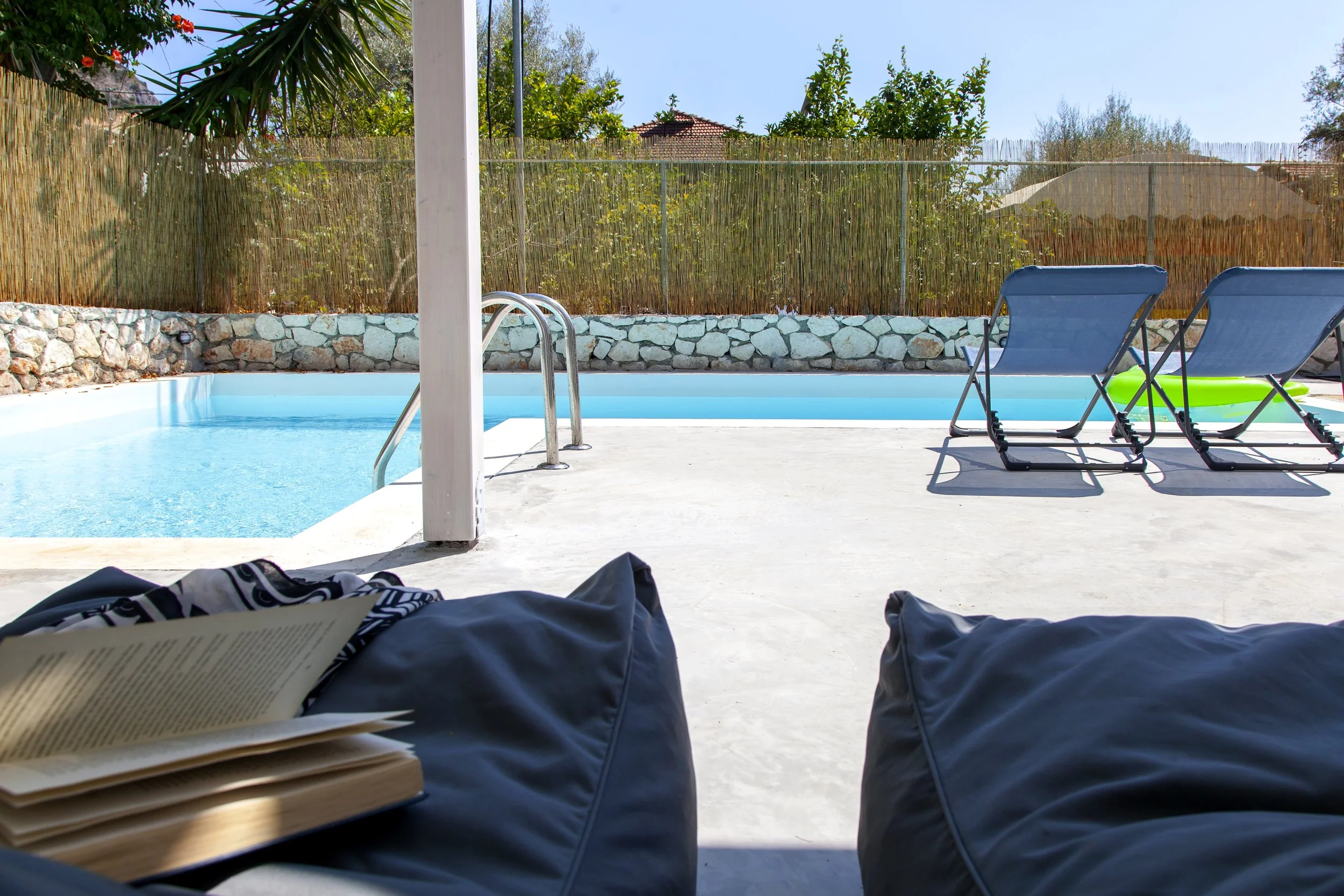 View from a person lying down with open book and clothing, looking at a backyard pool area with two lounge chairs, stone wall, bamboo fence, and trees under a clear blue sky.