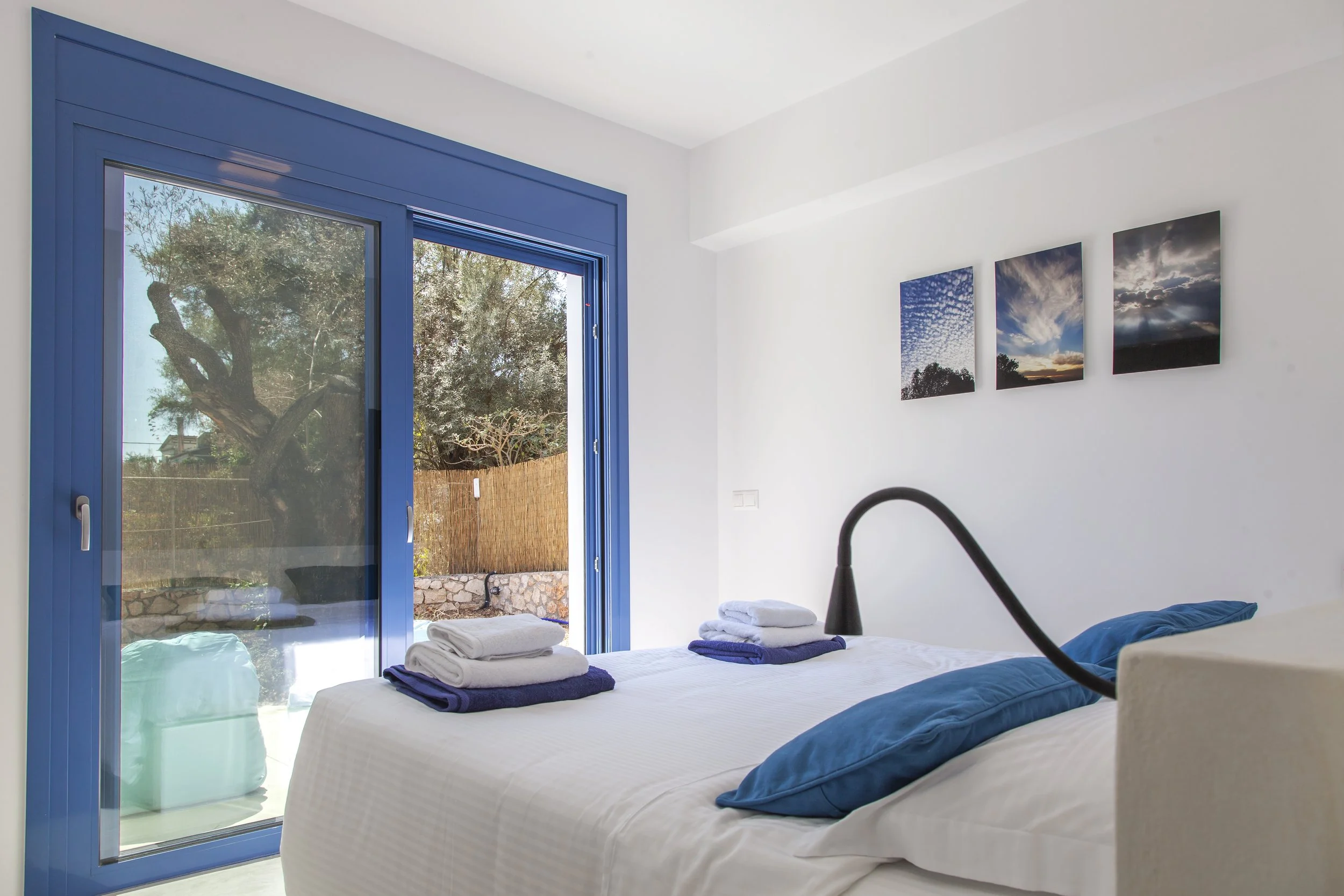 A bedroom with a large glass sliding door showing a view of trees outdoors. The bed has a white bedspread, with folded towels on top. There are three framed photographs of clouds and sky on the wall. The room has white walls and a black lamp on the b