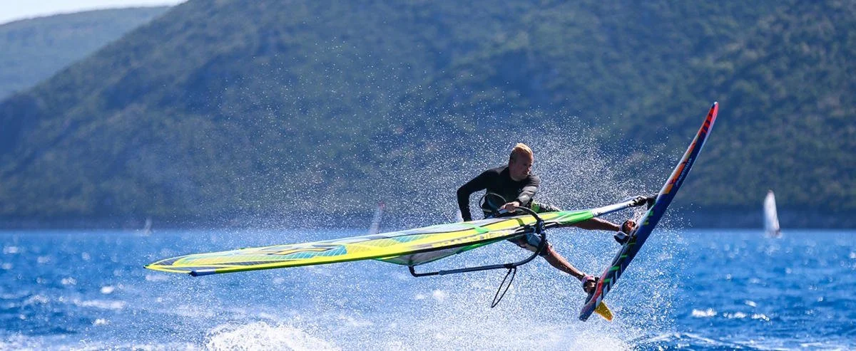 A person windsurfing on blue water with a mountainous background.