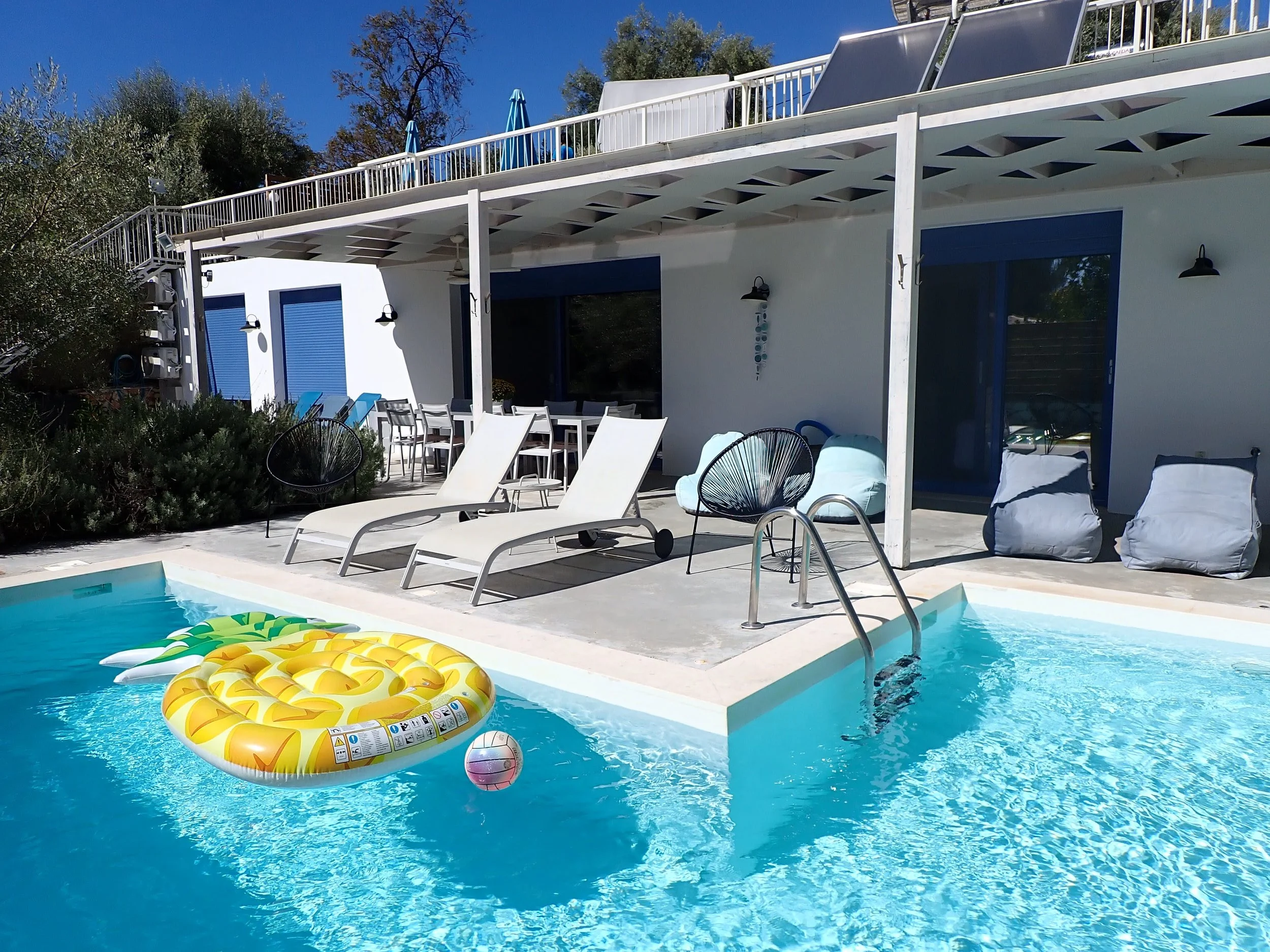 View of a backyard with a swimming pool, pool floats including a pineapple and a ball, lounge chairs, an outdoor dining table, a balcony with umbrellas, and a modern house with blue window frames under a clear blue sky.