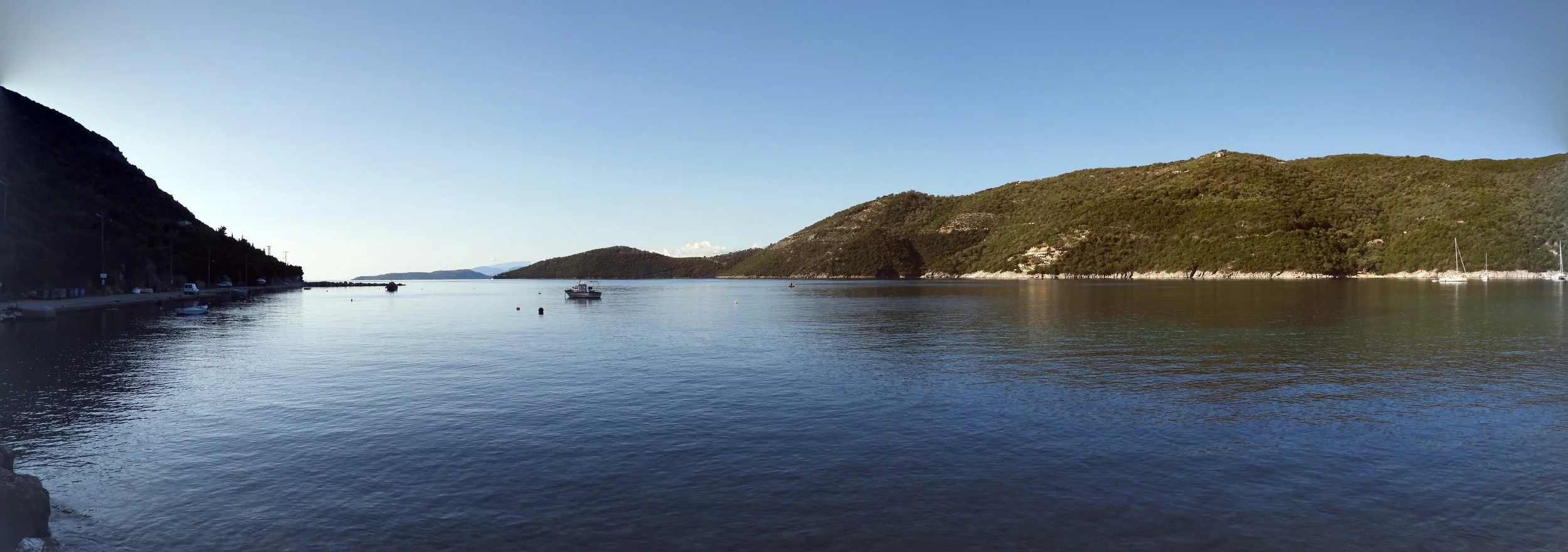 A calm body of water surrounded by green hills, with several small boats anchored and a few cars parked near the shoreline.