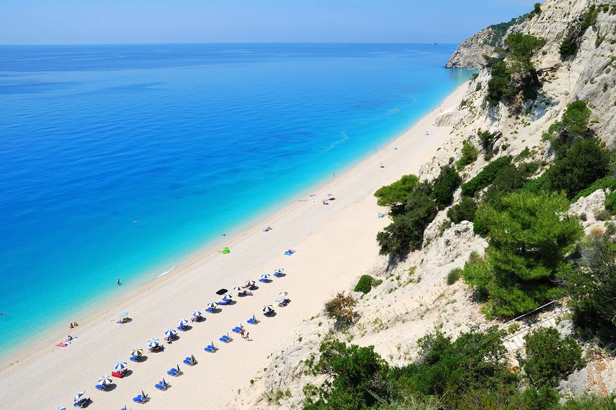 A sandy beach with colorful umbrellas and lounge chairs near clear, blue water, surrounded by green trees and white cliffs.