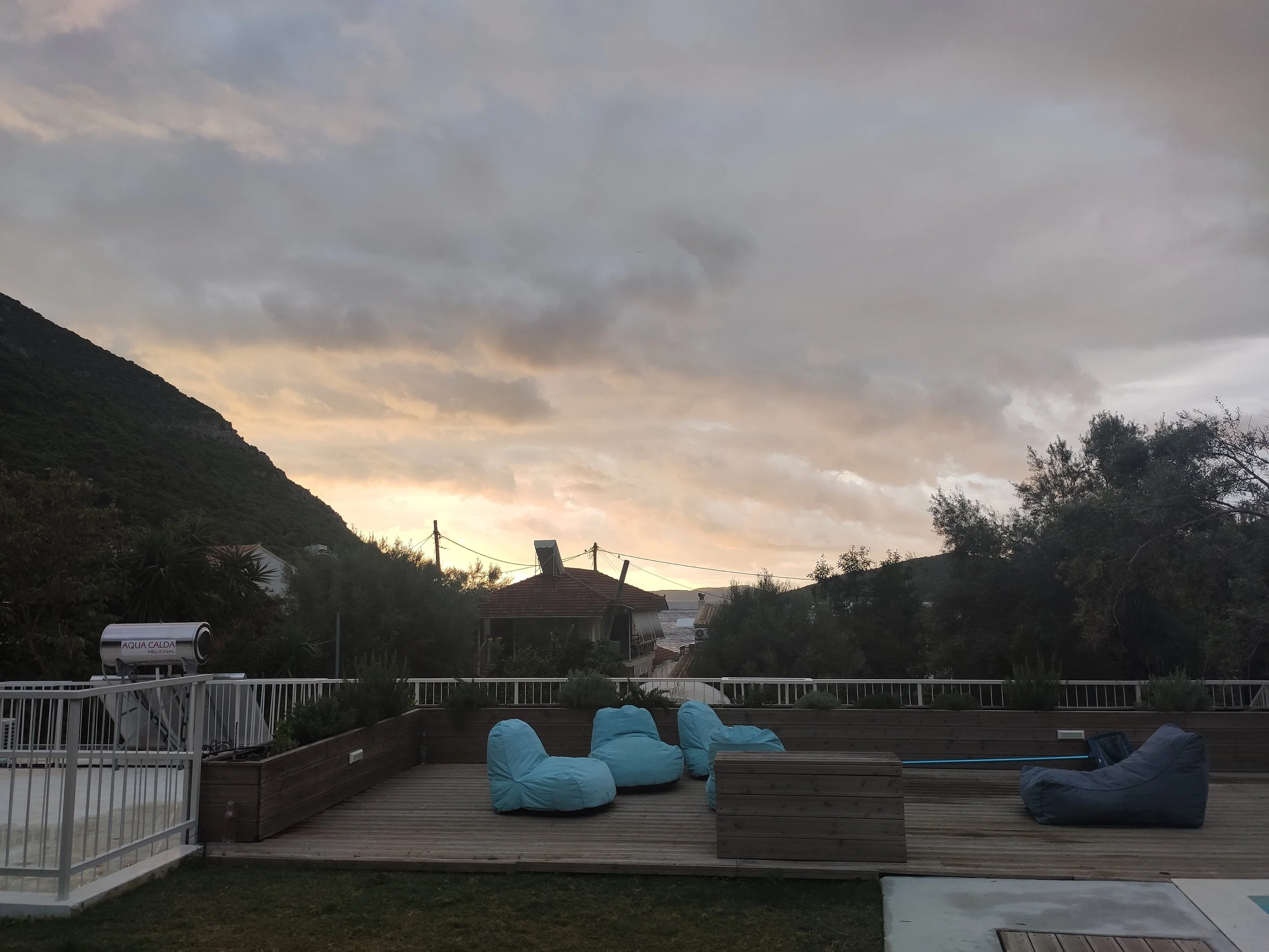A backyard patio with blue bean bag chairs, a wooden deck, and a white railing. The sky is cloudy with a hint of sunset in the distance, and there are trees and houses in the background, with mountains on the left side.