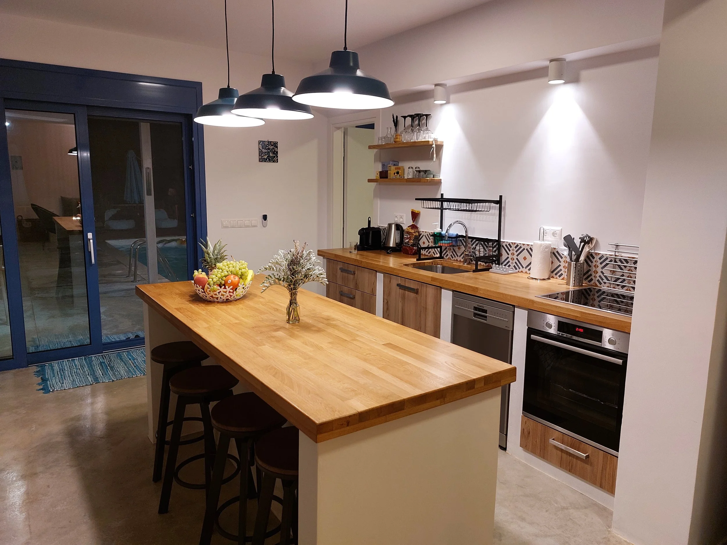 A modern kitchen with a wooden island, black pendant lights, white cabinetry, stainless steel appliances, and shelves with glassware and spices. There are outdoor sliding glass doors and a small rug visible.