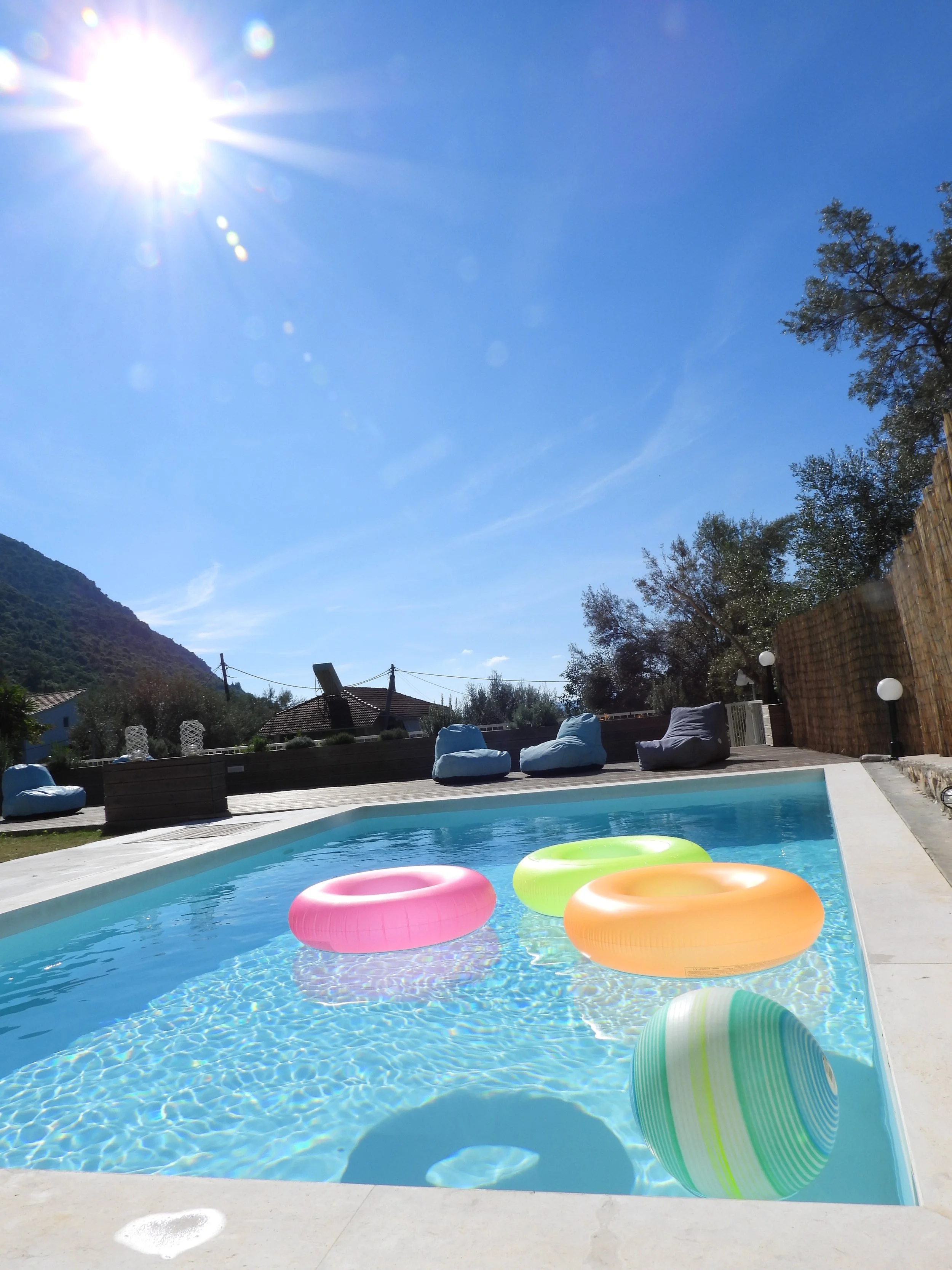 View of a backyard swimming pool with pastel-colored pool floaties, surrounded by outdoor seating, trees, a wooden fence, and a mountain in the background under a bright blue sky with a shining sun.