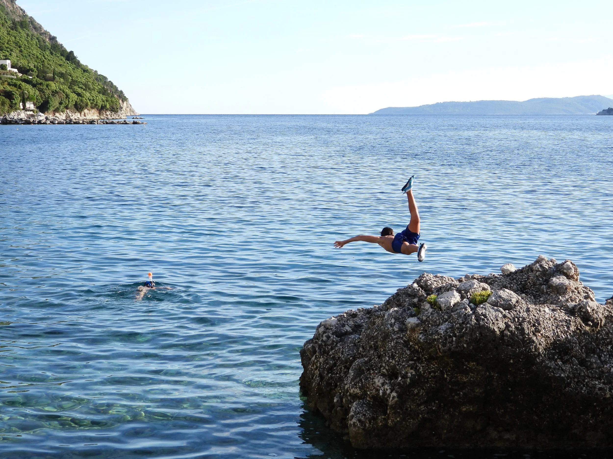 Person diving into the water near rocky shoreline by a calm, expansive body of water with green hills in the background.