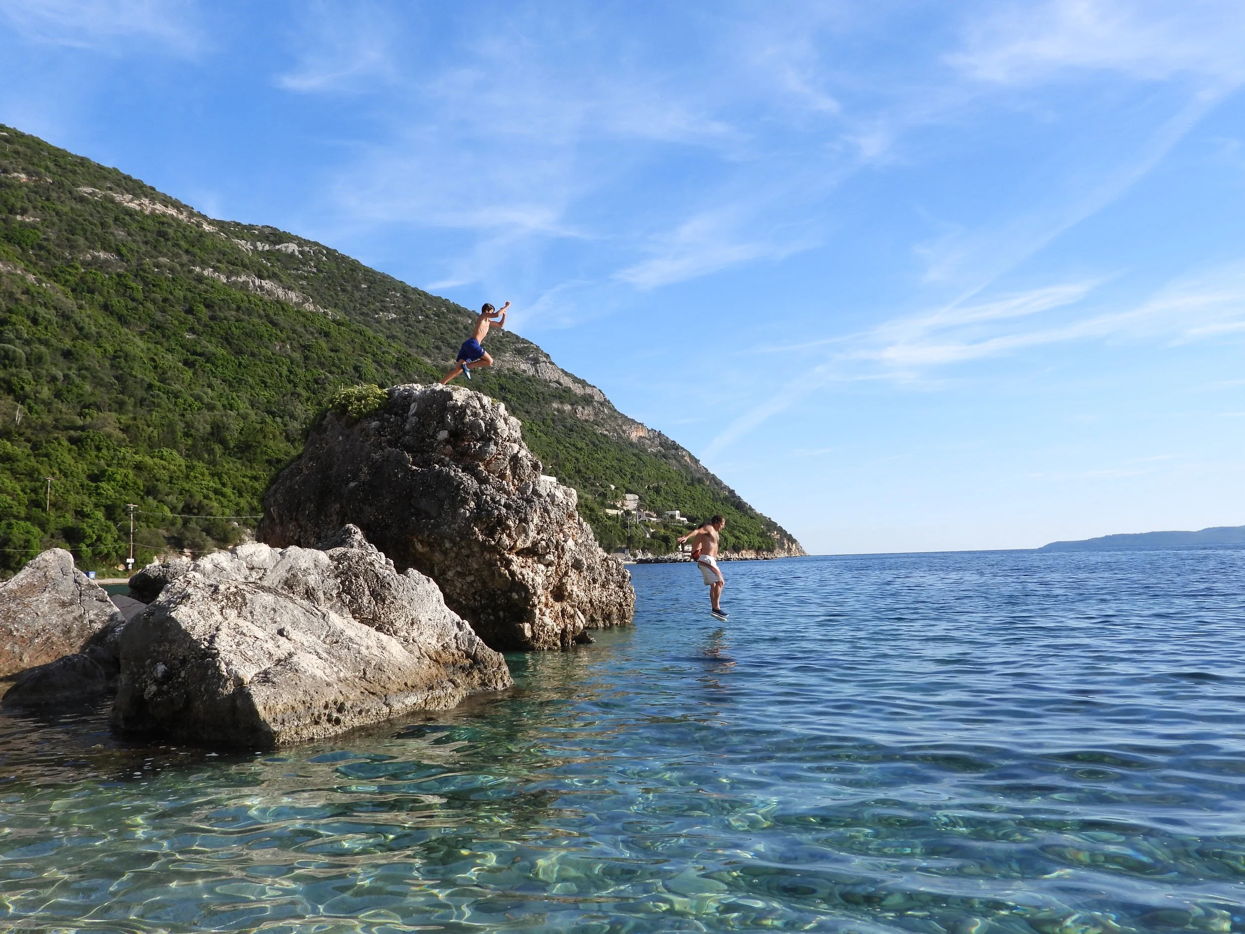 A child jumping off a rock into the water at a coastal area with a hillside covered in greenery and a clear blue sky.