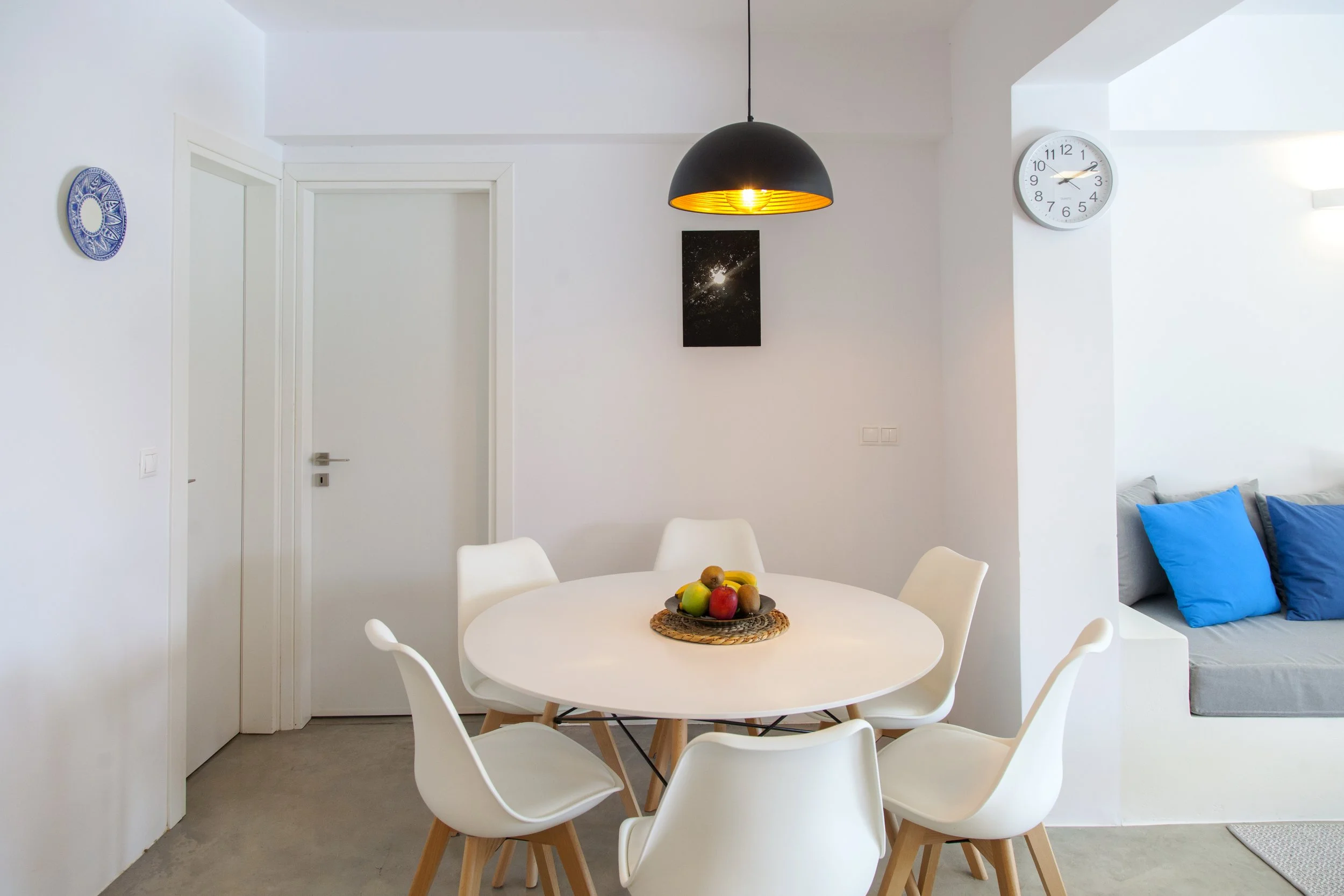A modern dining area featuring a round white table with six white chairs, a fruit bowl with apples and grapes, a black pendant light hanging from the ceiling, white walls with minimal decor including a wall clock and a framed picture, and part of a s
