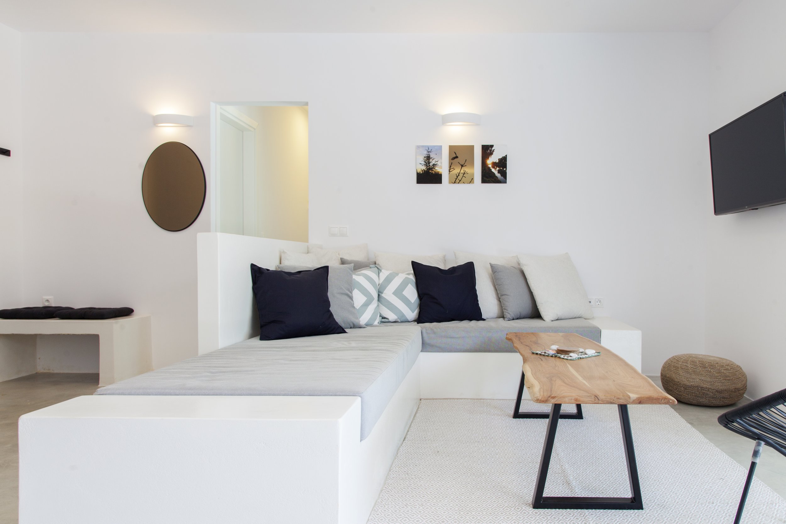 Modern living room with white sectional sofa, black and white pillows, wall art of nature scenes, wall-mounted lights, a wooden coffee table, and a television on the wall.