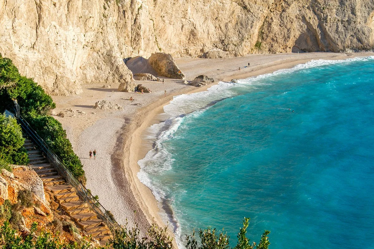 A beach with sandy shoreline, turquoise water, rocky cliffs, and a staircase leading down to the beach, with some people sunbathing and walking.