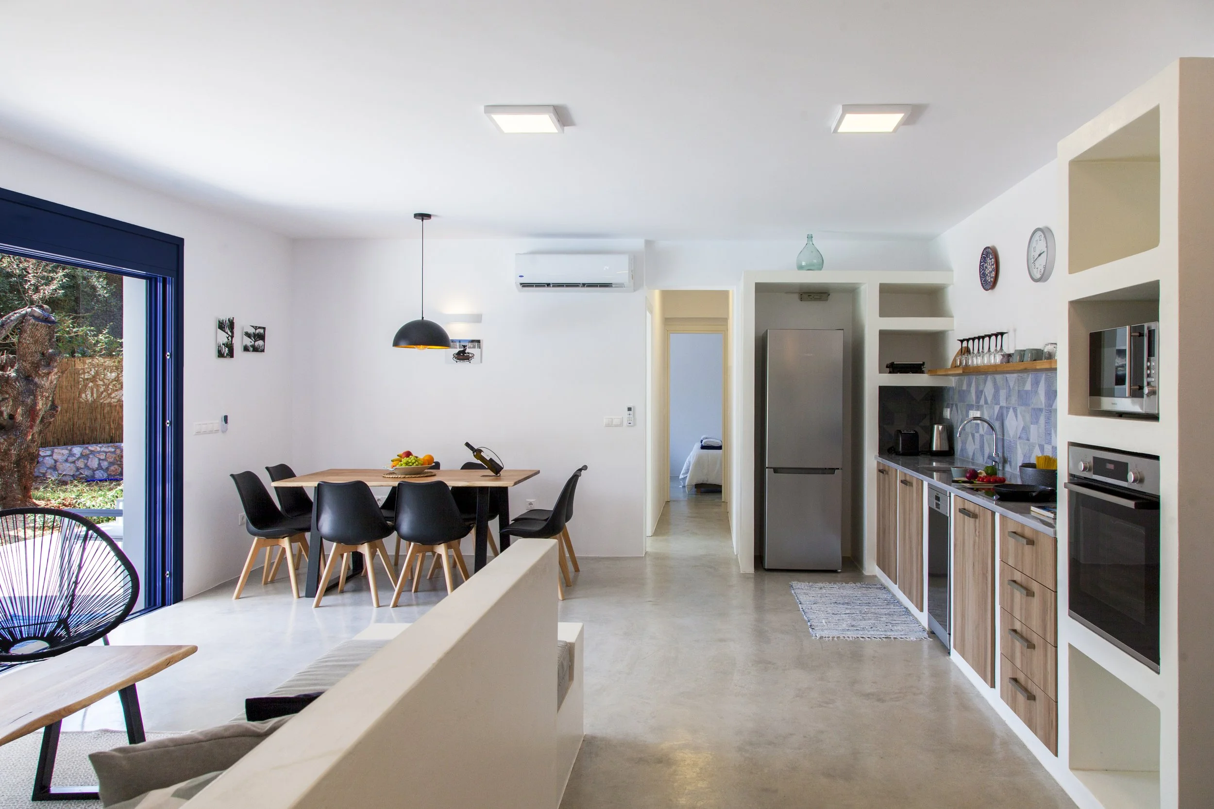 Interior of a modern kitchen and dining area with white walls, wooden cabinets, stainless steel appliances, a black dining table, and black chairs. Large sliding door with a view of trees outside.