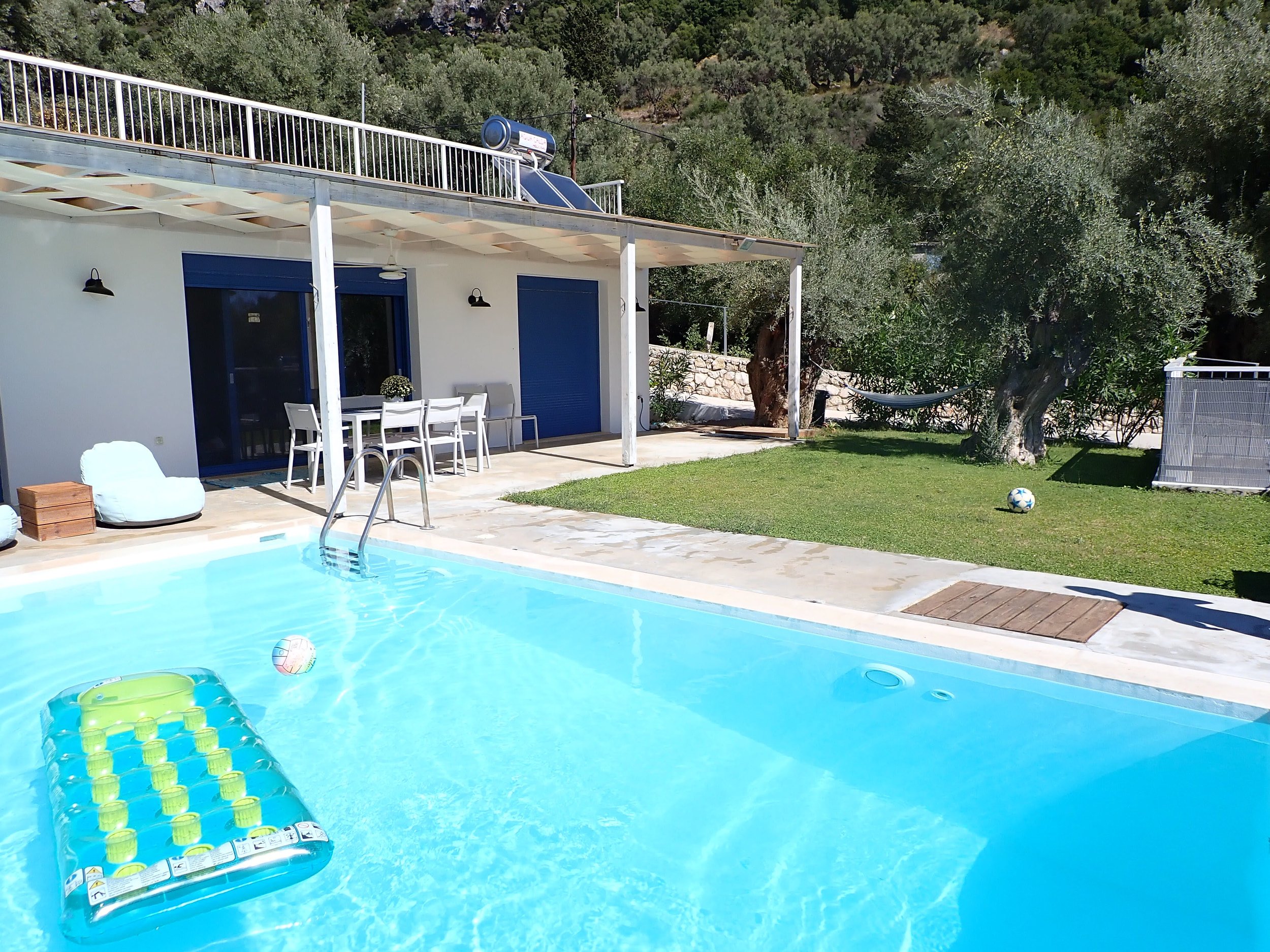 Swimming pool with floating pool toys, patio furniture, and a house with a balcony and solar panel on the roof, surrounded by trees and greenery.