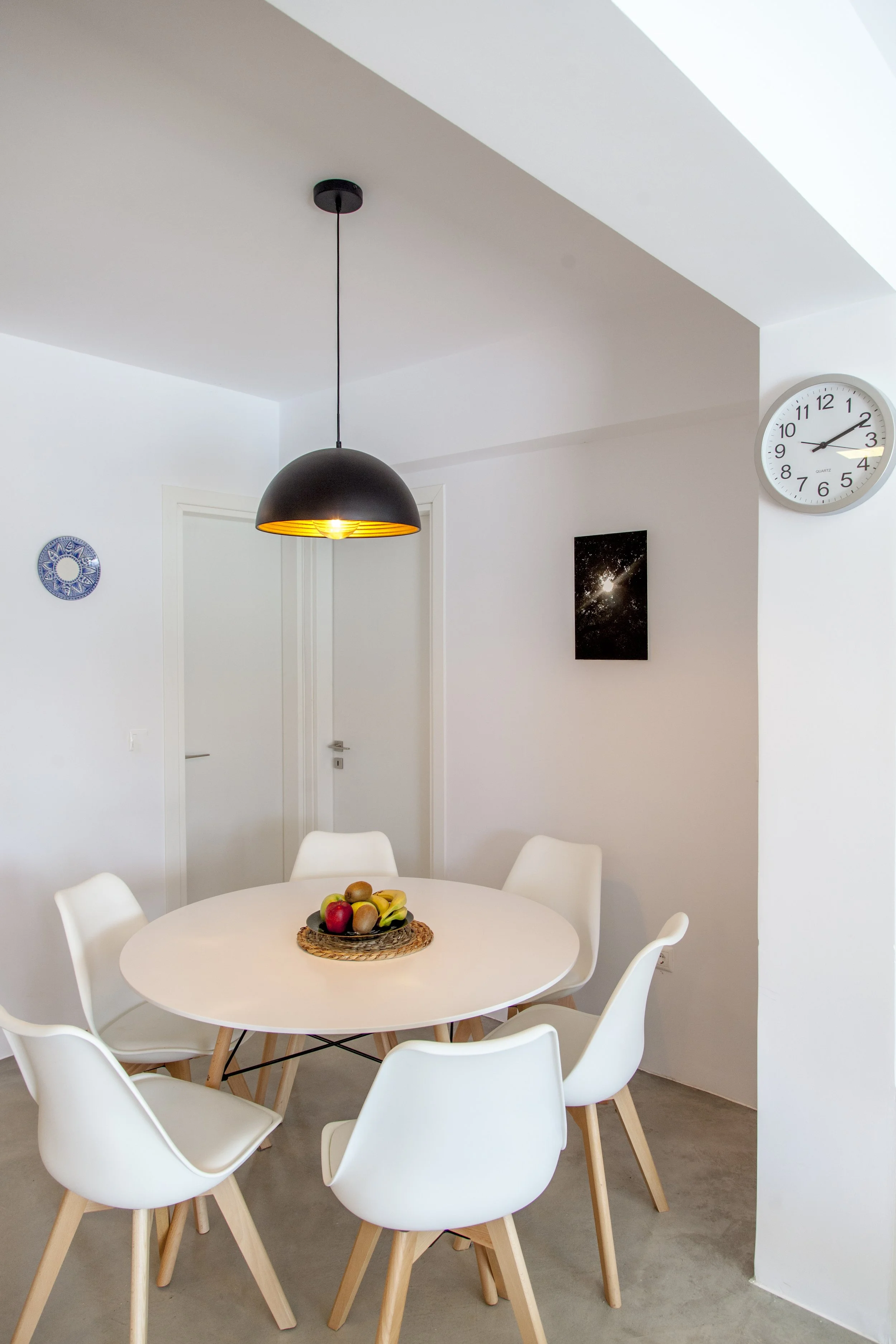 A modern dining area with a round white table surrounded by white chairs with wooden legs. A basket of fresh fruit, including apples, kiwis, and bananas, is placed on the table. A black pendant light hangs from the ceiling above the table, turned on.