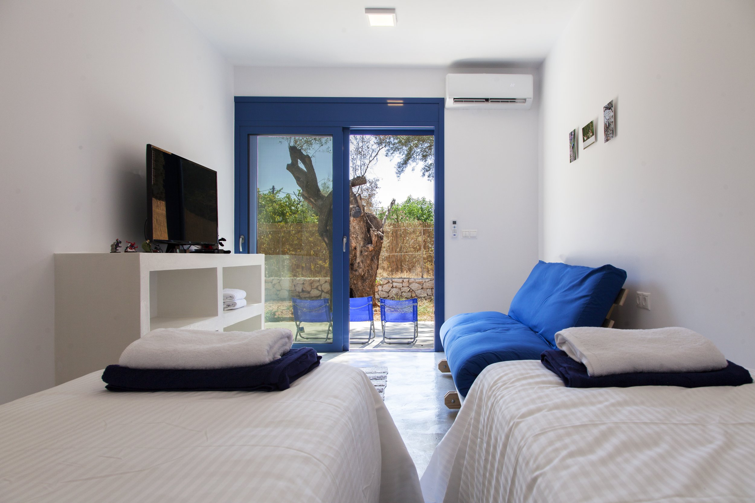 A bright, minimalist living room with two beds covered in white striped bedding, a blue sofa, a TV on a white shelf, and a sliding glass door leading to a patio with outdoor chairs and a tree.