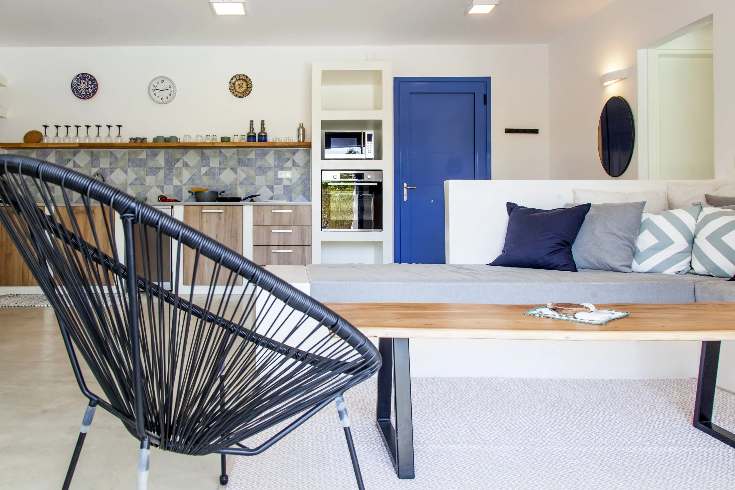 A modern open-plan living space with a kitchen area in the background featuring a microwave and oven built into a white shelving unit. The kitchen has a geometric-patterned tile backsplash and a wooden countertop with various glasses and bottles. In 
