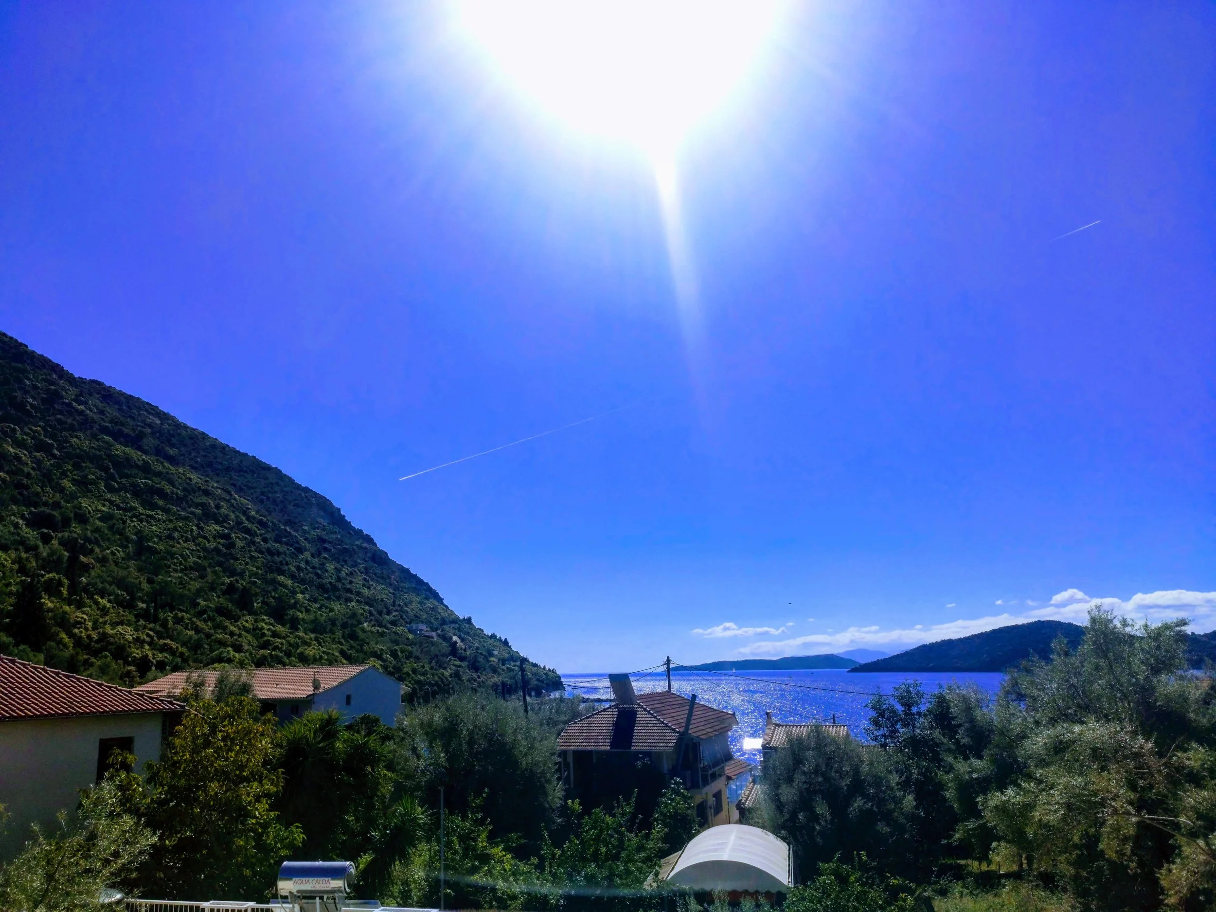 A bright sunny day over a coastal village with houses, green trees, a mountain, and a body of water in the background.