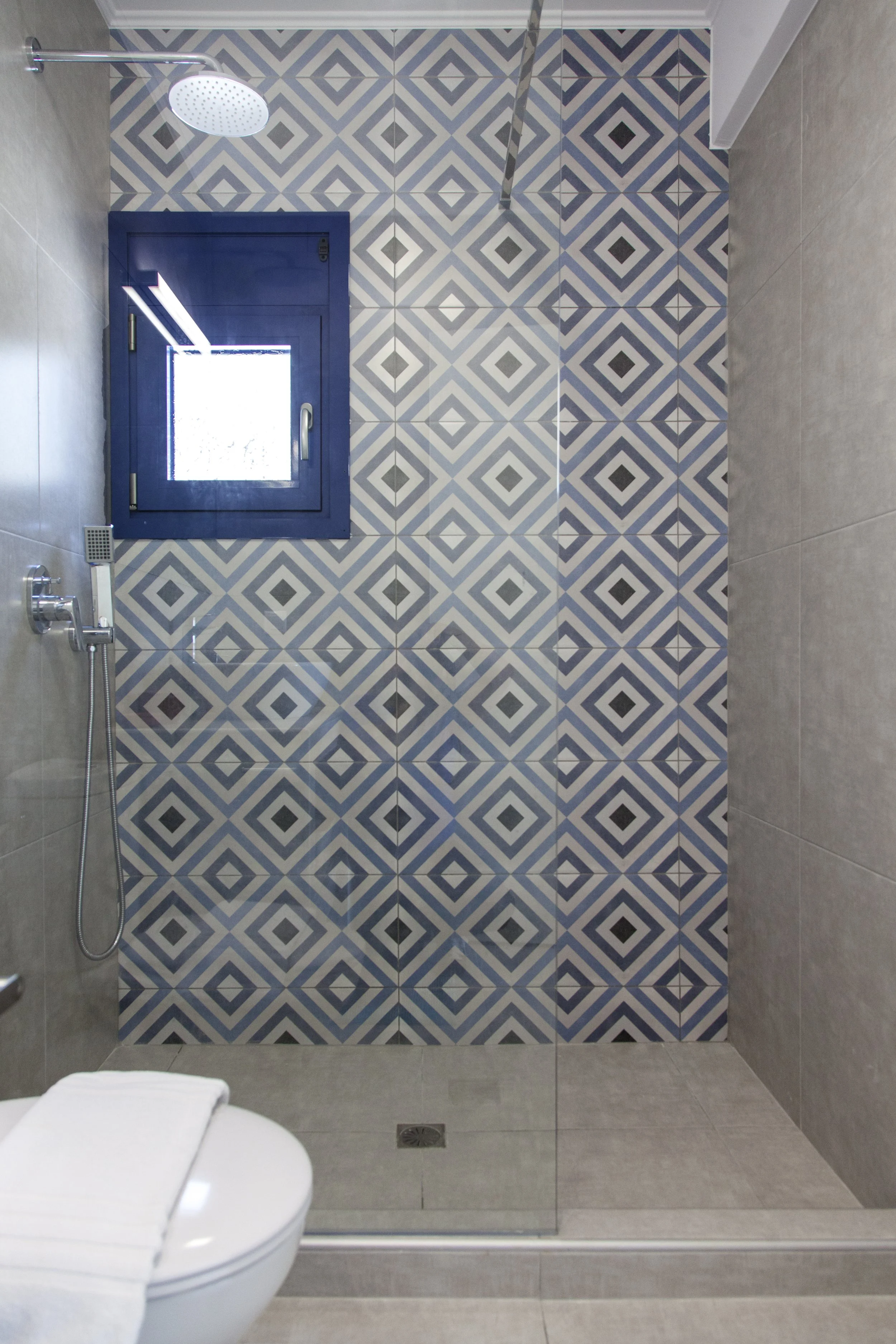 Modern bathroom with a geometric-patterned tile wall, a glass shower door, a rainfall showerhead, and a white toilet.