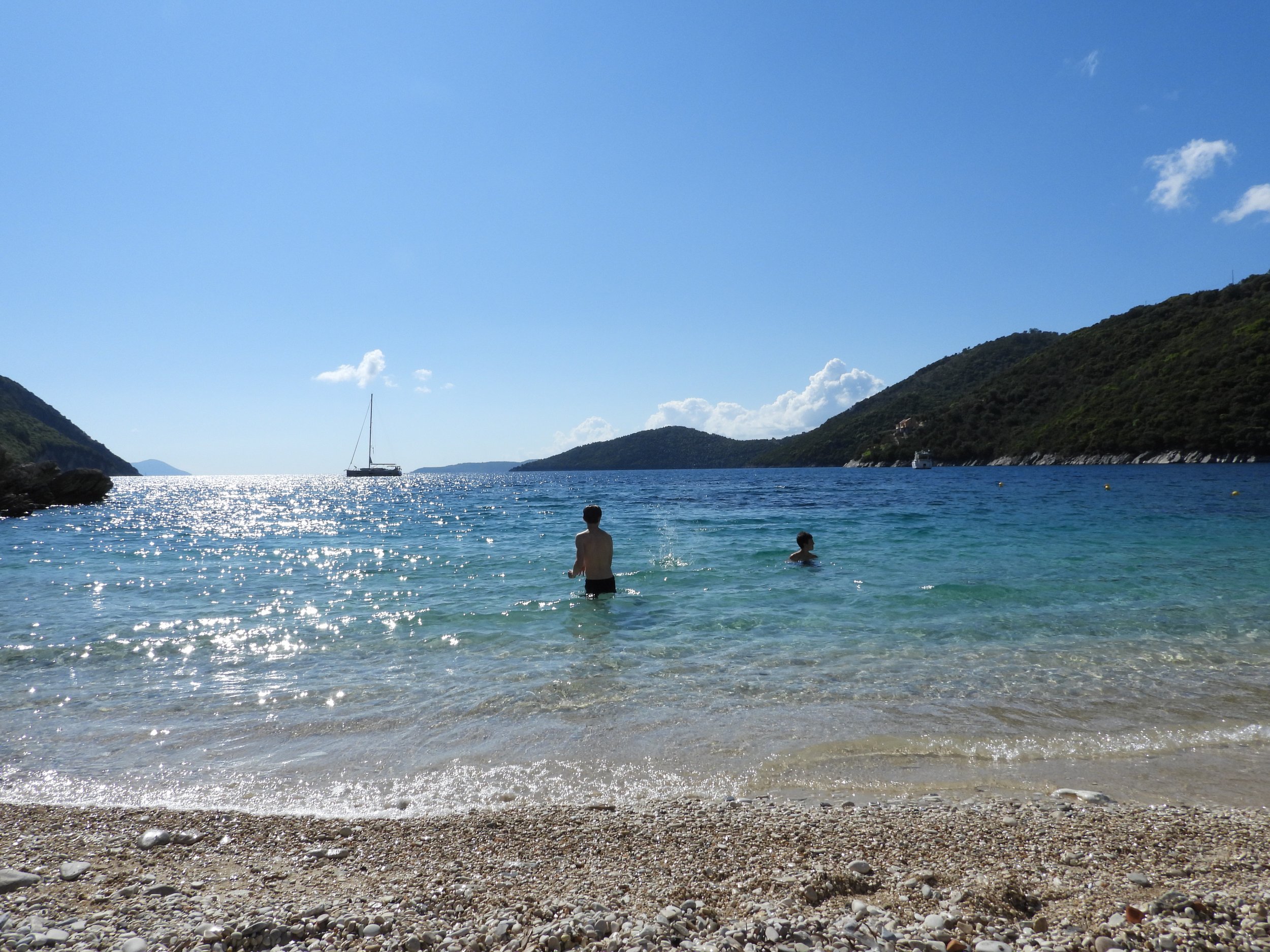 Clear blue ocean water with two people wading in the shallow part of the beach, surrounded by green hills and a sailboat in the distance under a bright sunny sky.
